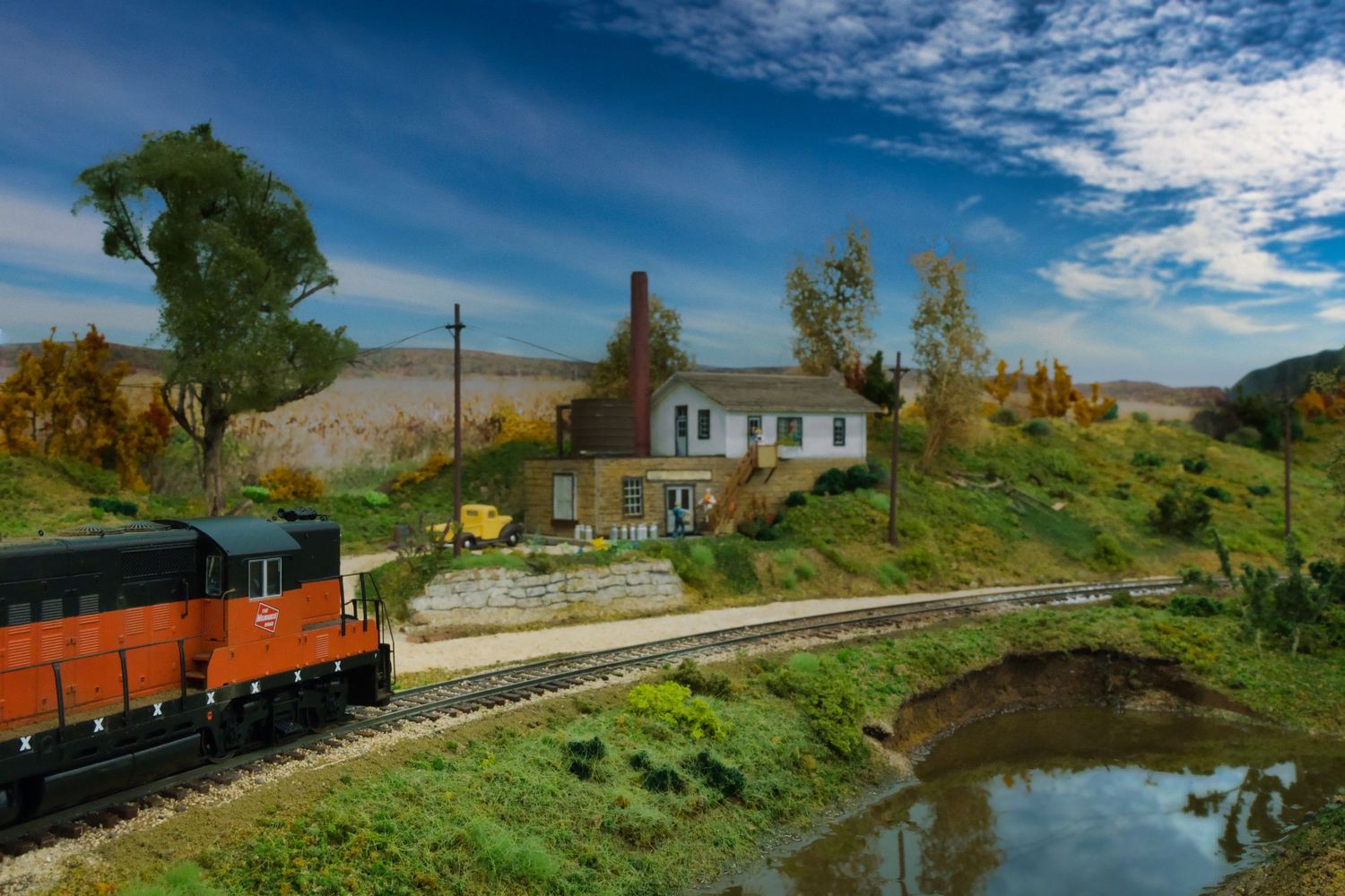 Model train layout featuring miniature landscapes with tracks, buildings, and a scenic pond under dramatic blue skies.