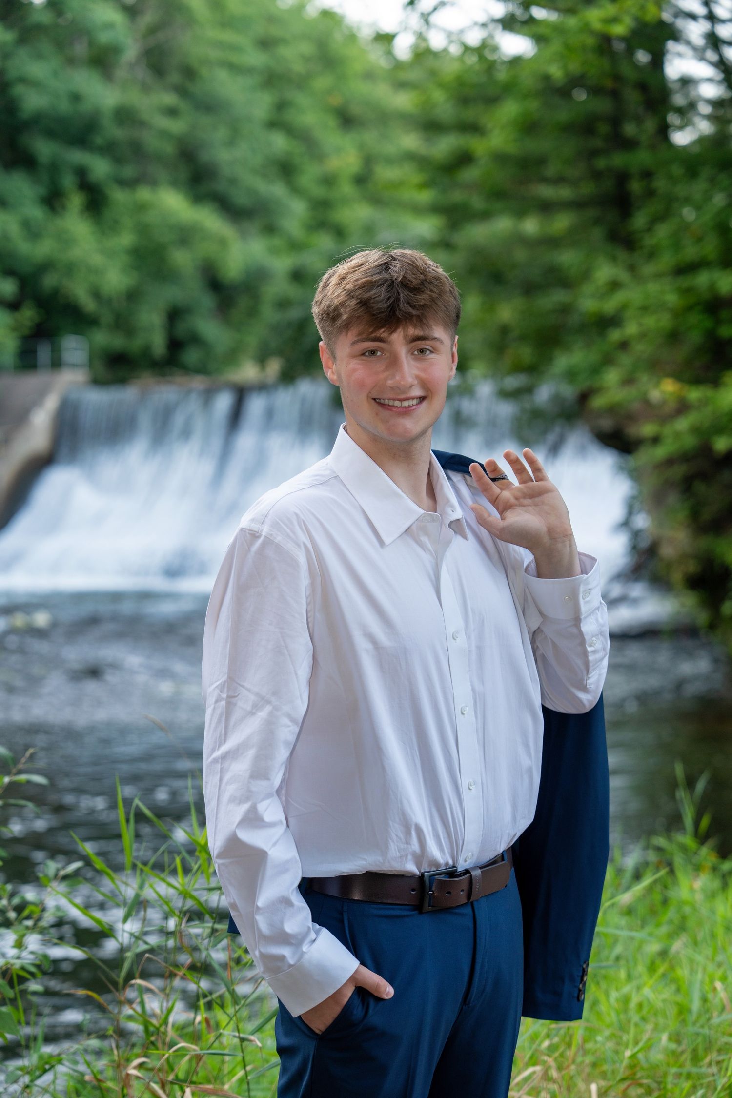 A person in a white dress shirt and navy pants poses in front of a cascading waterfall on a summer day.
