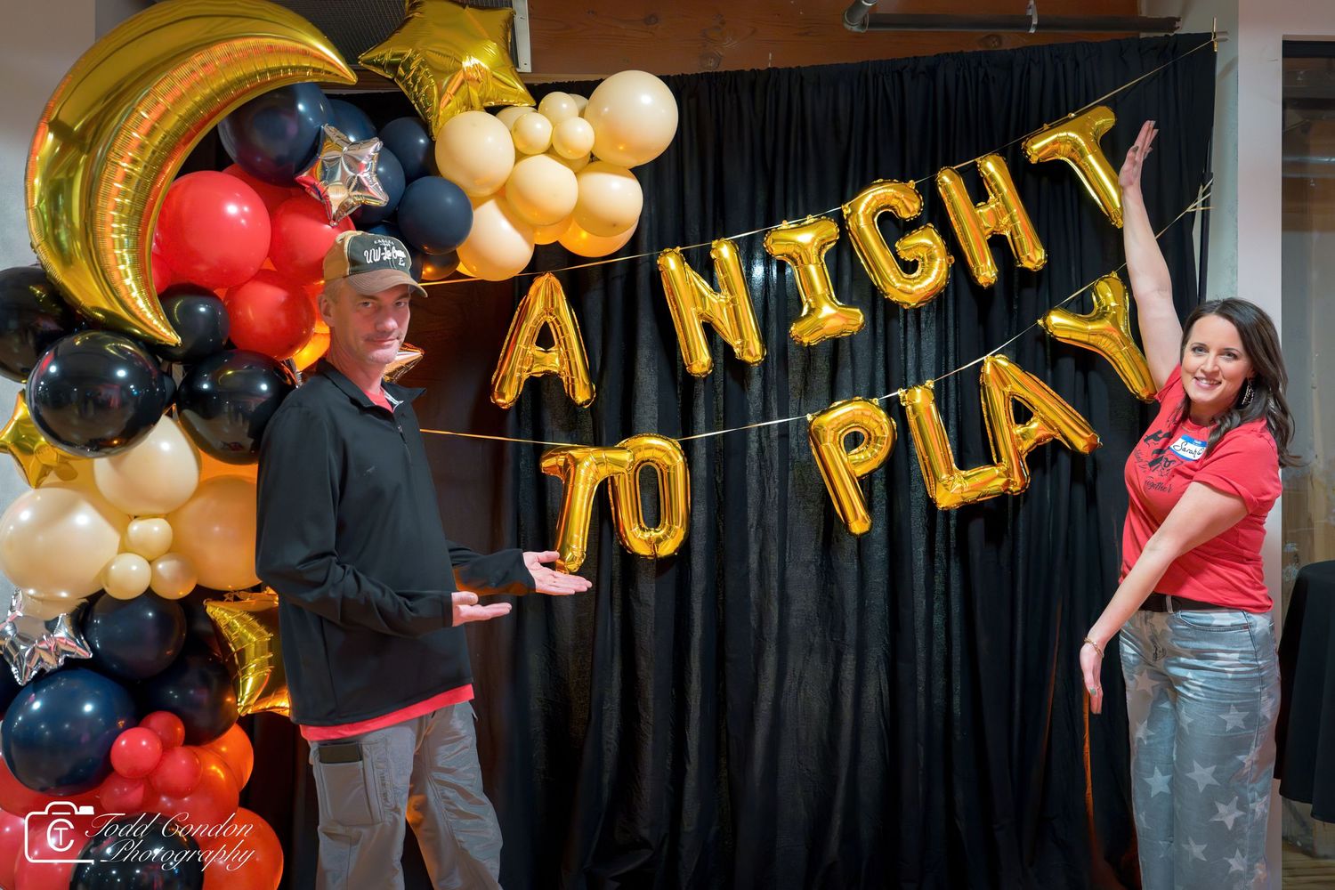 Two people pose beside gold balloon letters spelling A Night To Play on a black backdrop with colorful balloon decorations.