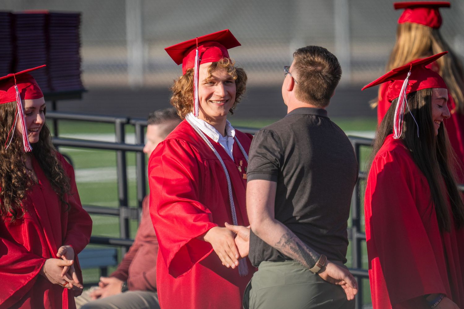 High school graduate in red cap and gown shaking hands at graduation ceremony outdoors.