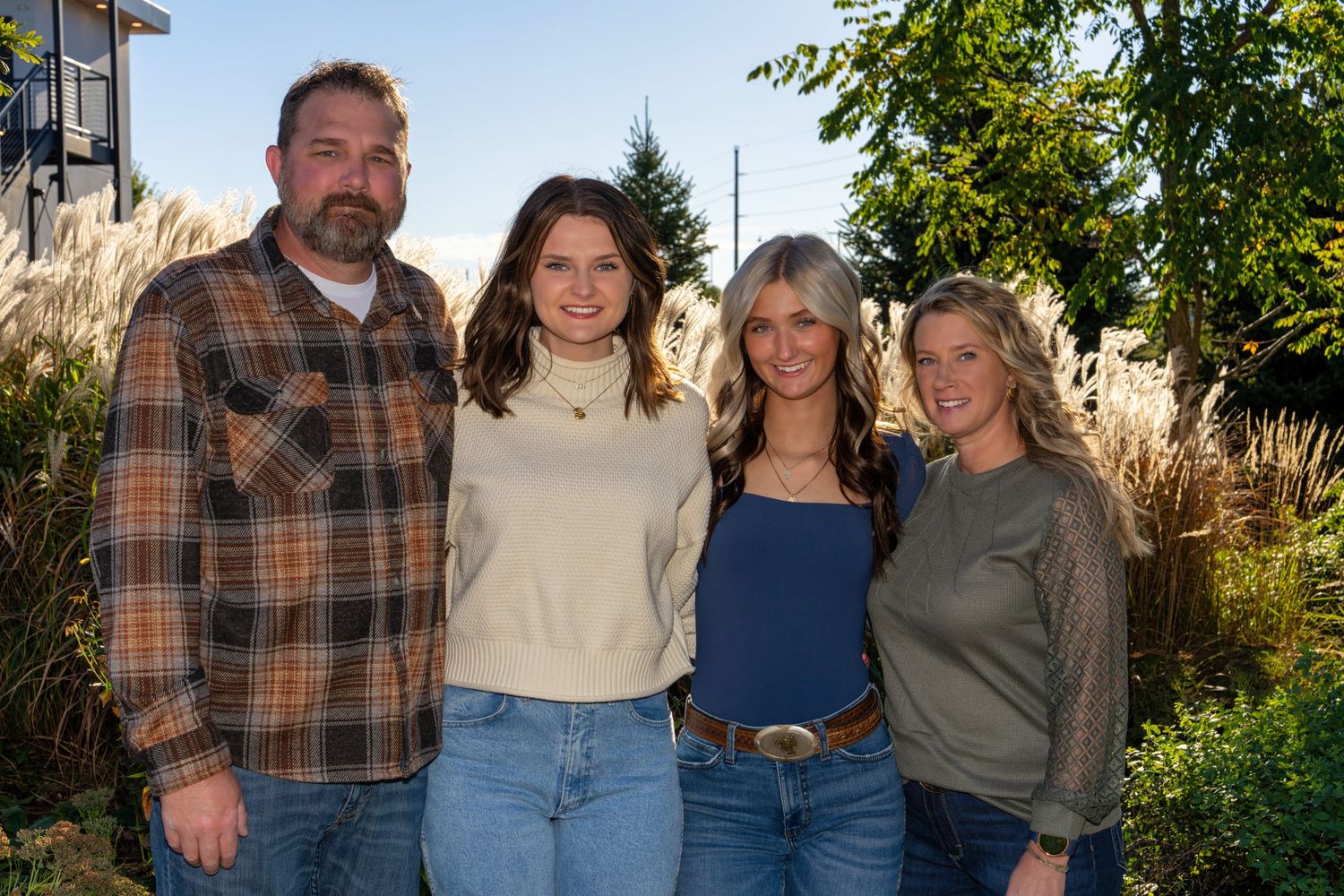 Four smiling adults posing together outdoors in casual attire surrounded by lush greenery and ornamental grasses.