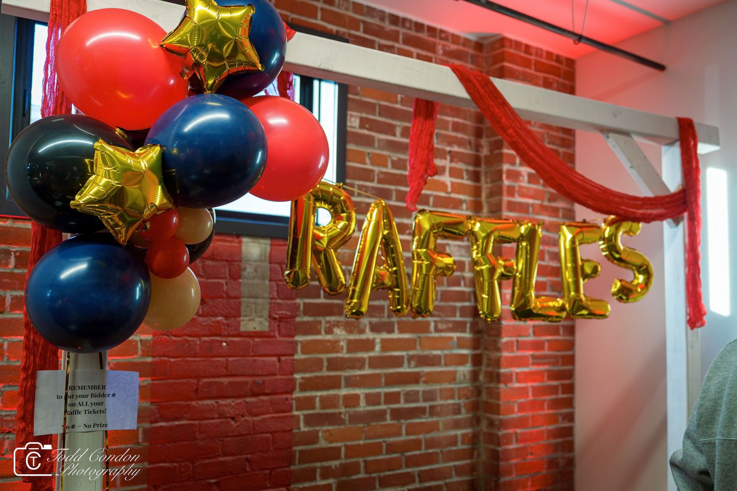 Gold letter balloon sign spelling RAFFLES with red, blue, and gold balloons against a brick wall.