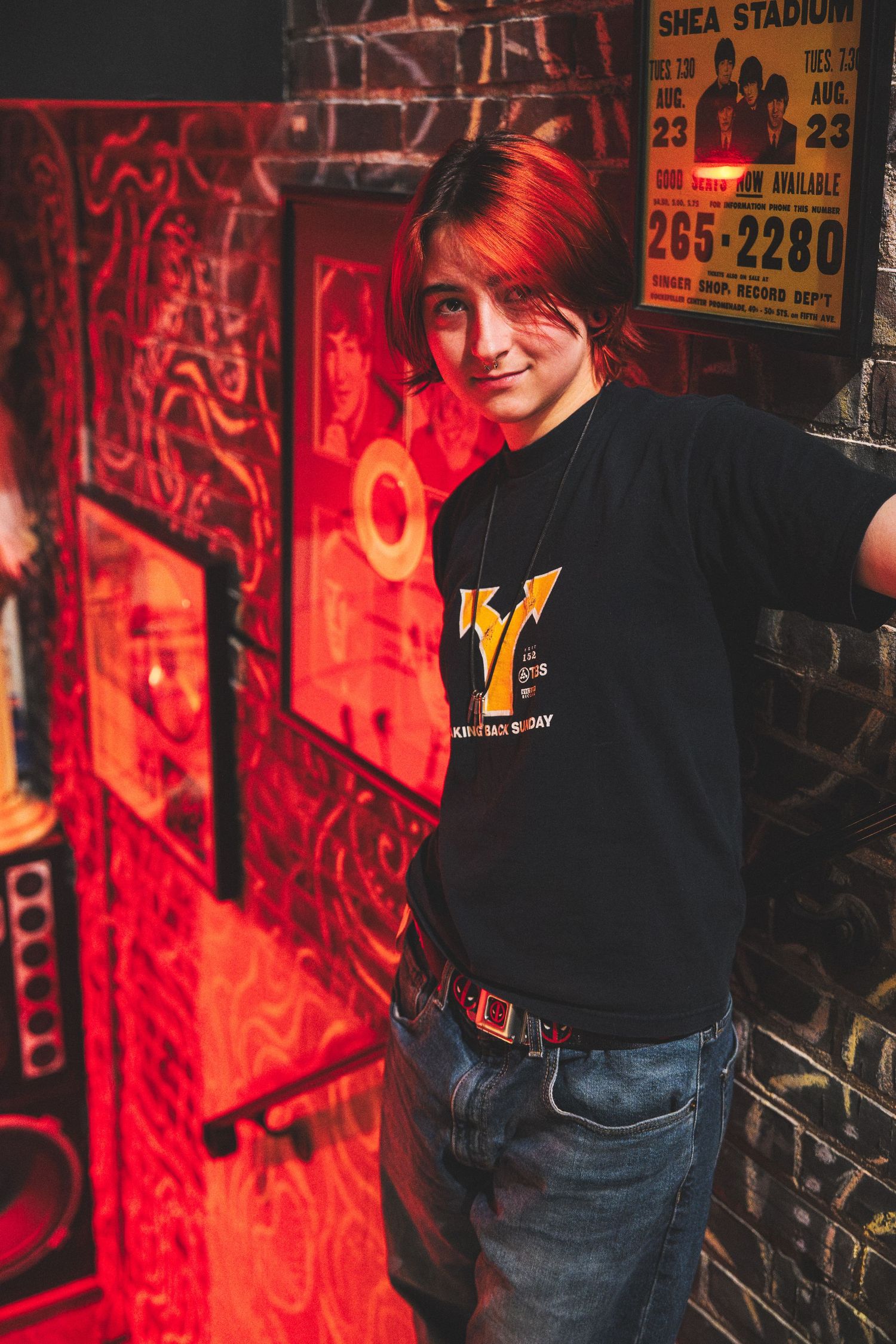 A person with vibrant red hair wearing a black t-shirt and jeans stands against a wall with red neon lighting.