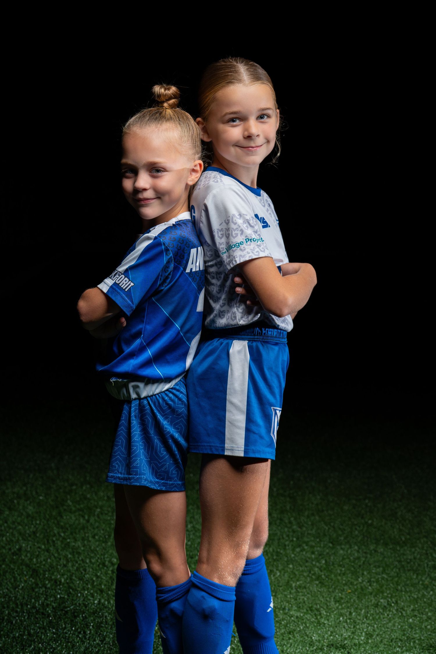 Two young soccer players in blue uniforms share a friendly hug on a green turf field.