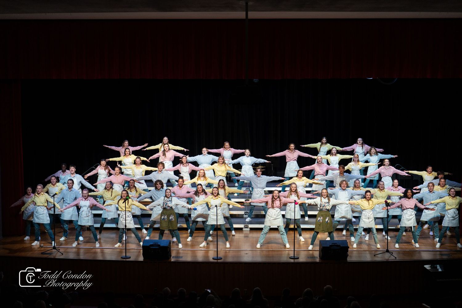 Large group of dancers in light blue and white costumes perform synchronized routine on dark stage during show.