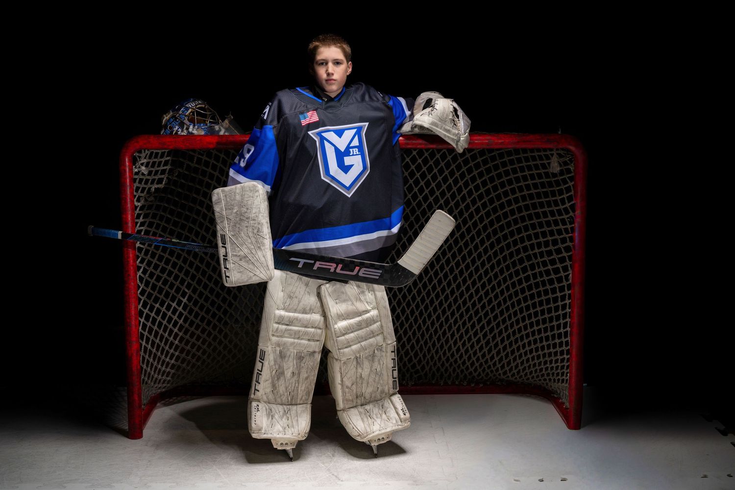 A hockey goalie in blue and white uniform makes a dramatic save in front of a black backdrop and red goal net.