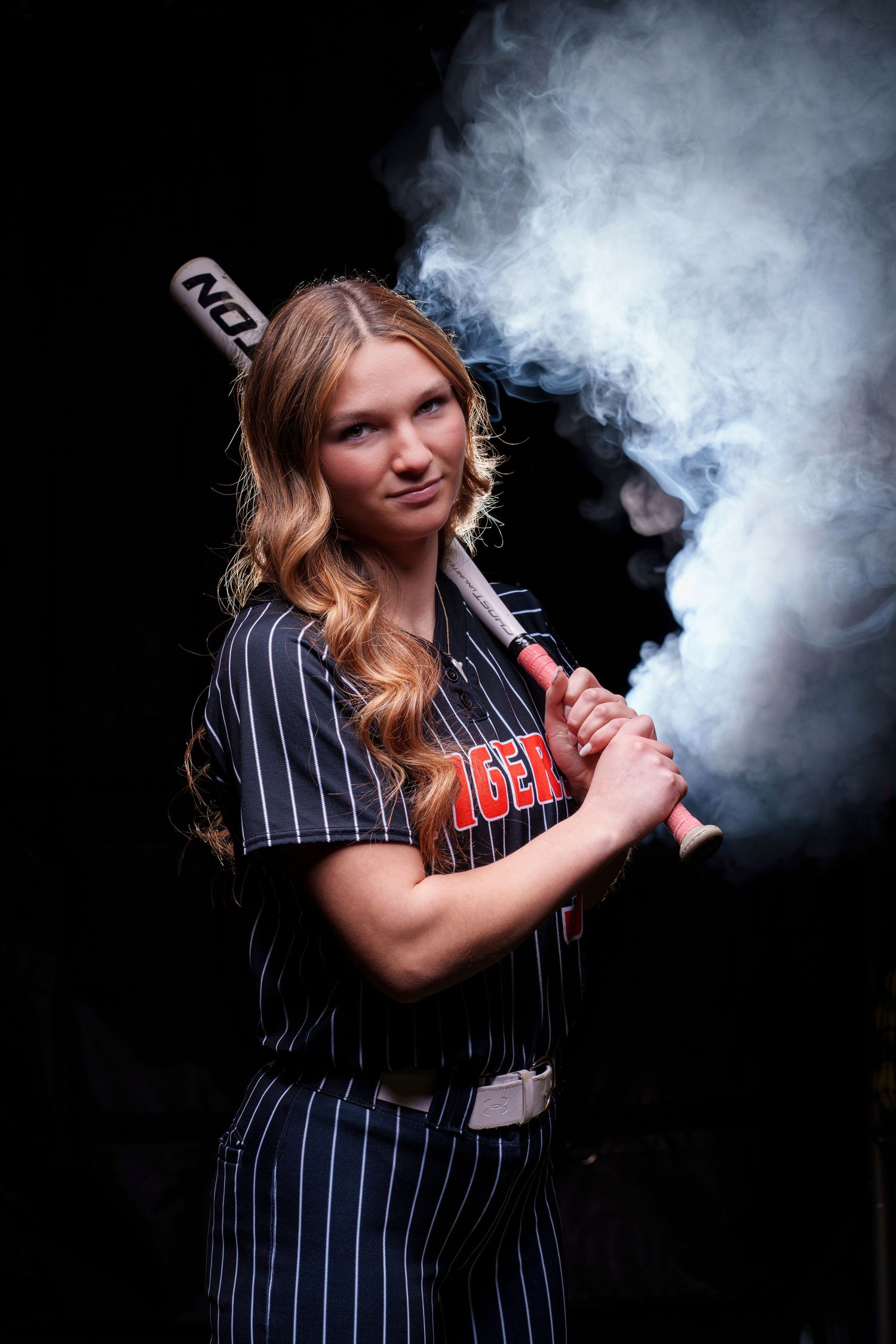 Young female softball player in pinstripe uniform holding a bat with dramatic smoke effect on dark background.