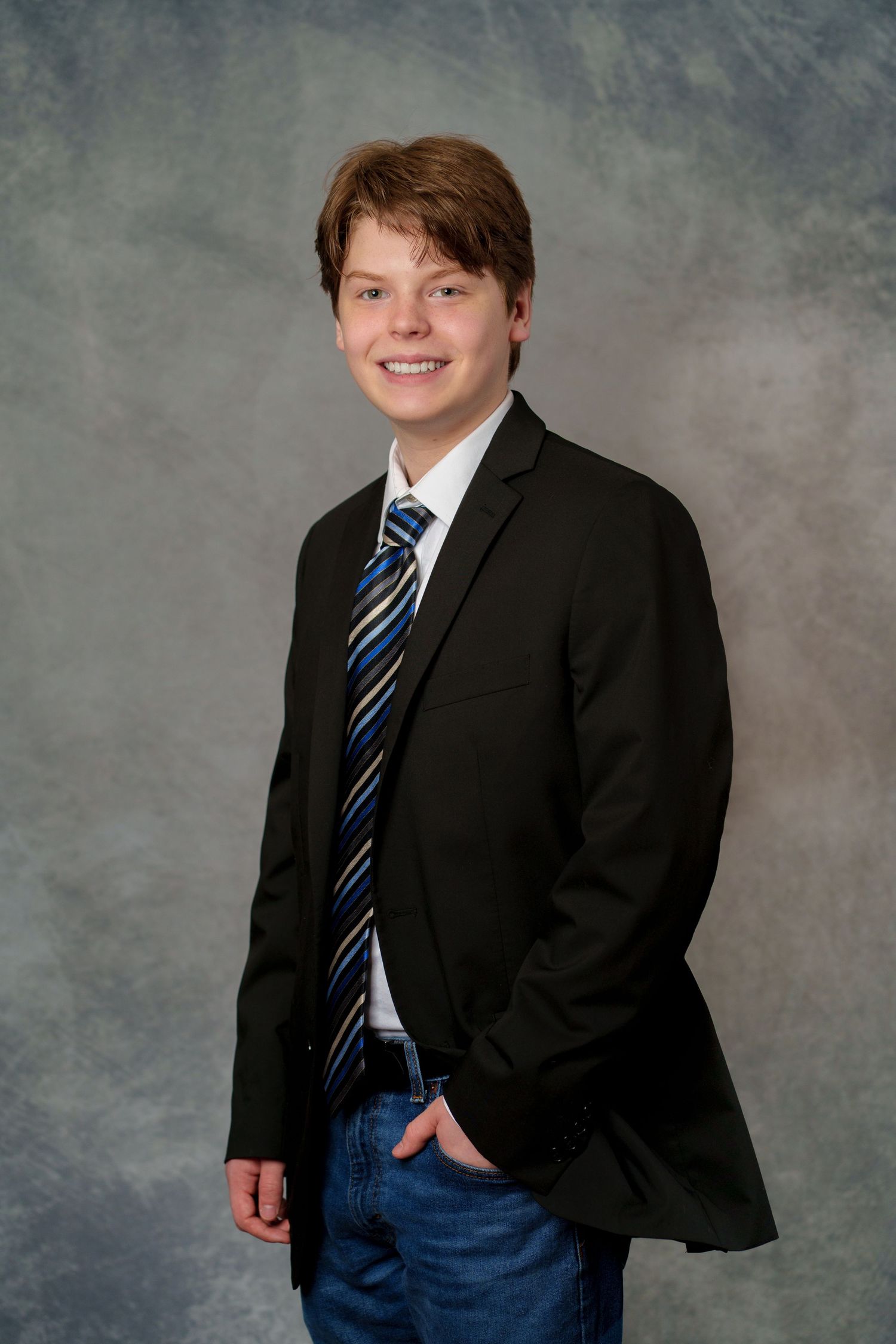 Smiling teenage boy in black blazer, striped tie, and jeans poses for a formal portrait photo.