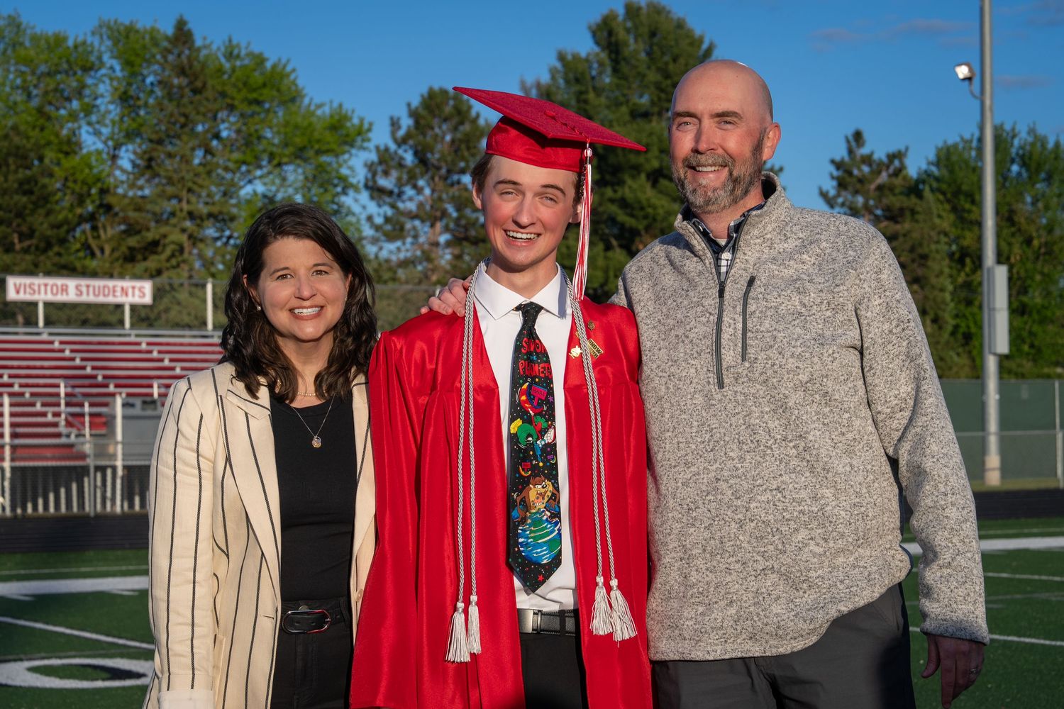 High school graduate in red cap and gown poses with proud parents on football field at graduation ceremony.