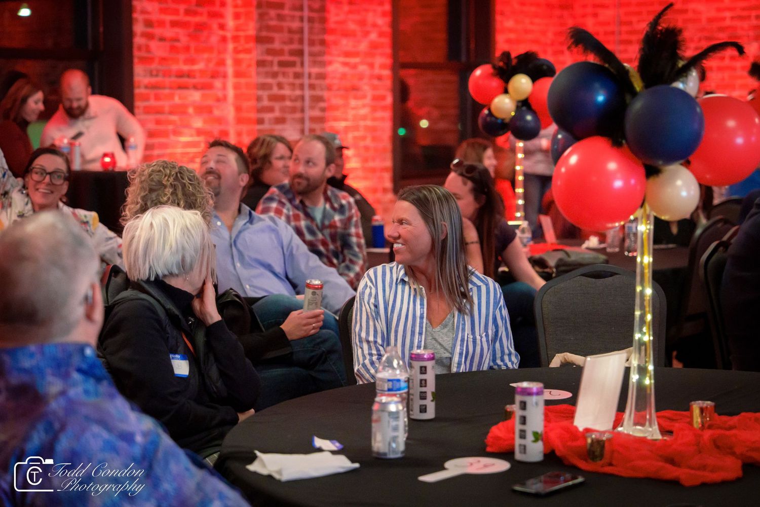 Guests laughing and socializing at a festive event with colorful balloon centerpieces and exposed brick walls in the background.
