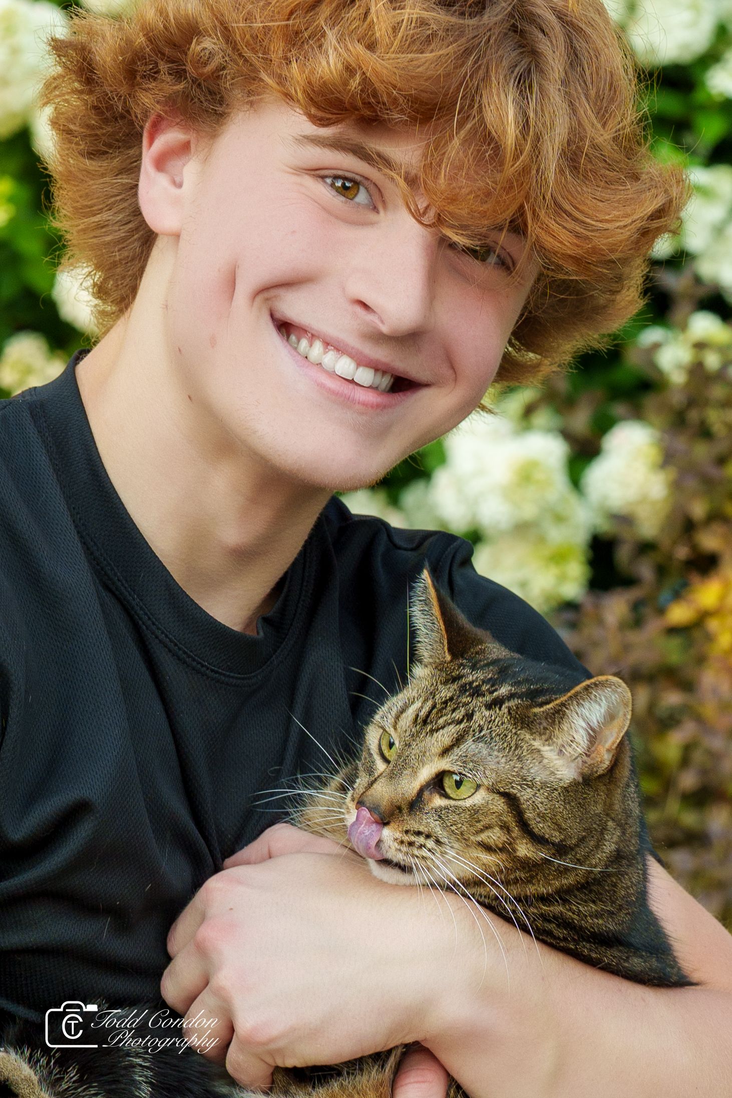 Smiling teenage boy with curly red hair holding a tabby cat in front of white flowers.