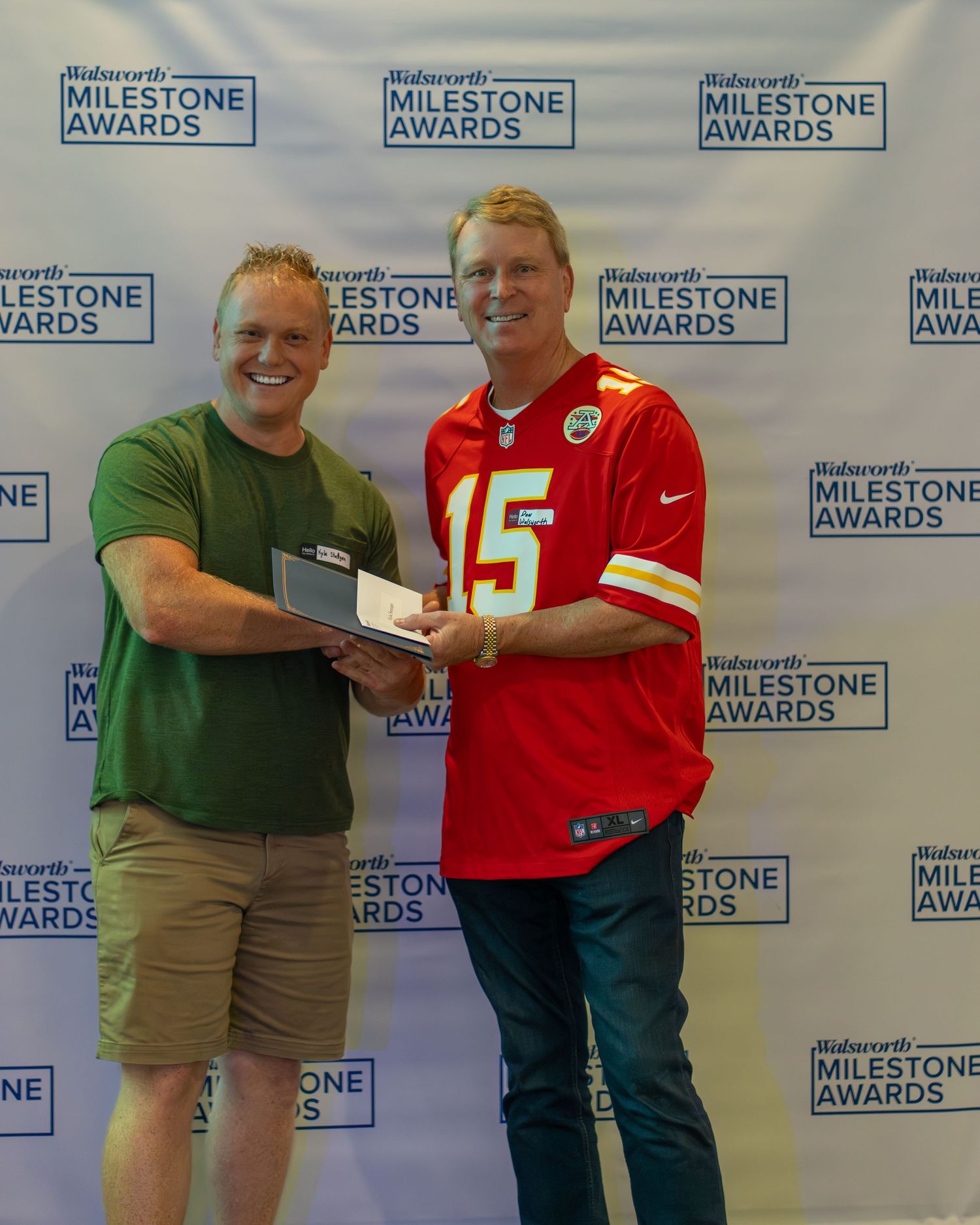 Two people pose together at a Milestone Awards event backdrop with one wearing a Kansas City Chiefs jersey number 15.