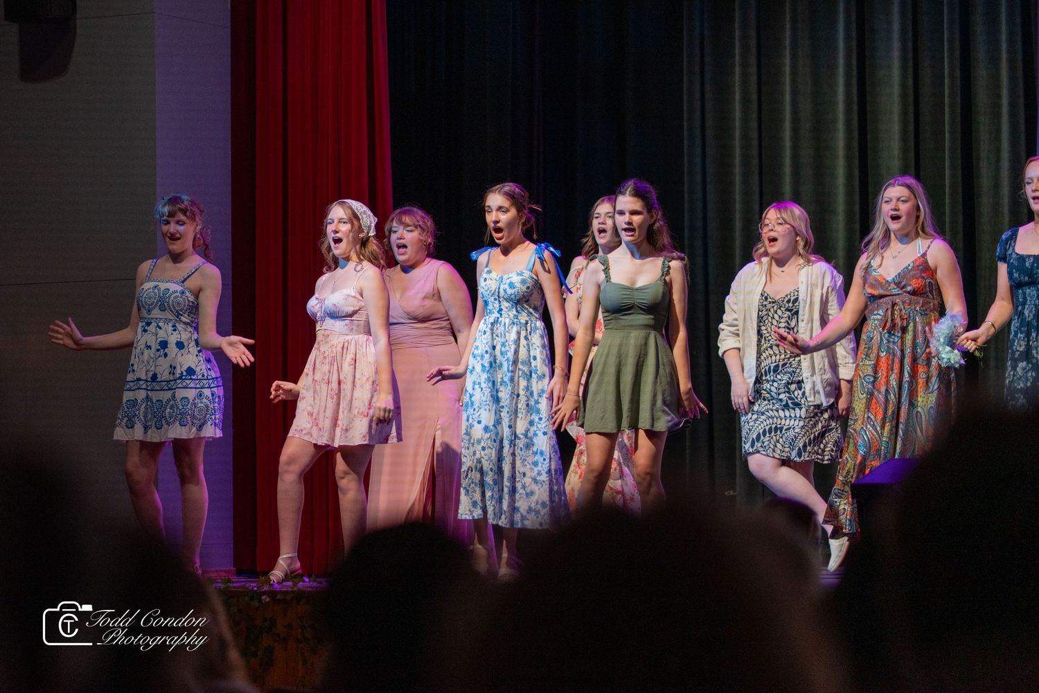 Performers in colorful floral dresses stand together on a theater stage during a musical performance.