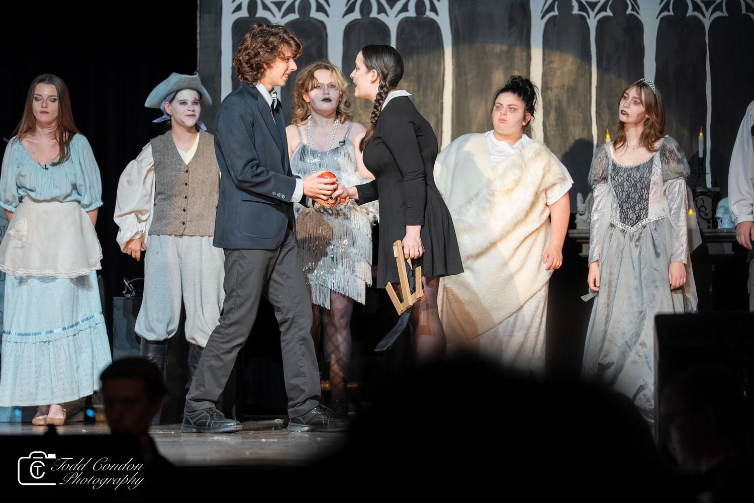 Performers in period costumes stand on stage during a theatrical production with ornate iron gates in the background.