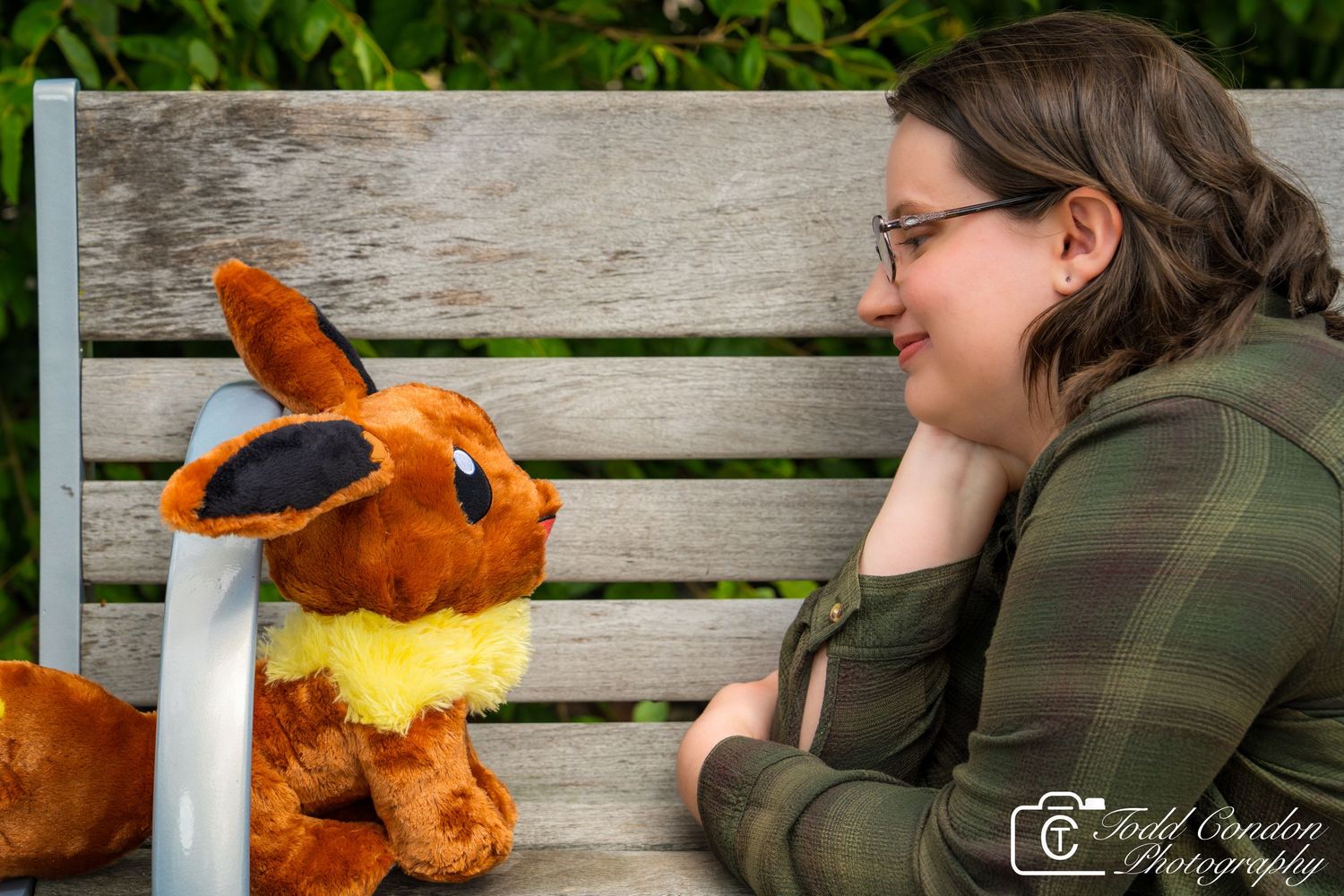 A plush Eevee Pokemon toy sits on a wooden bench next to someone wearing a green jacket.