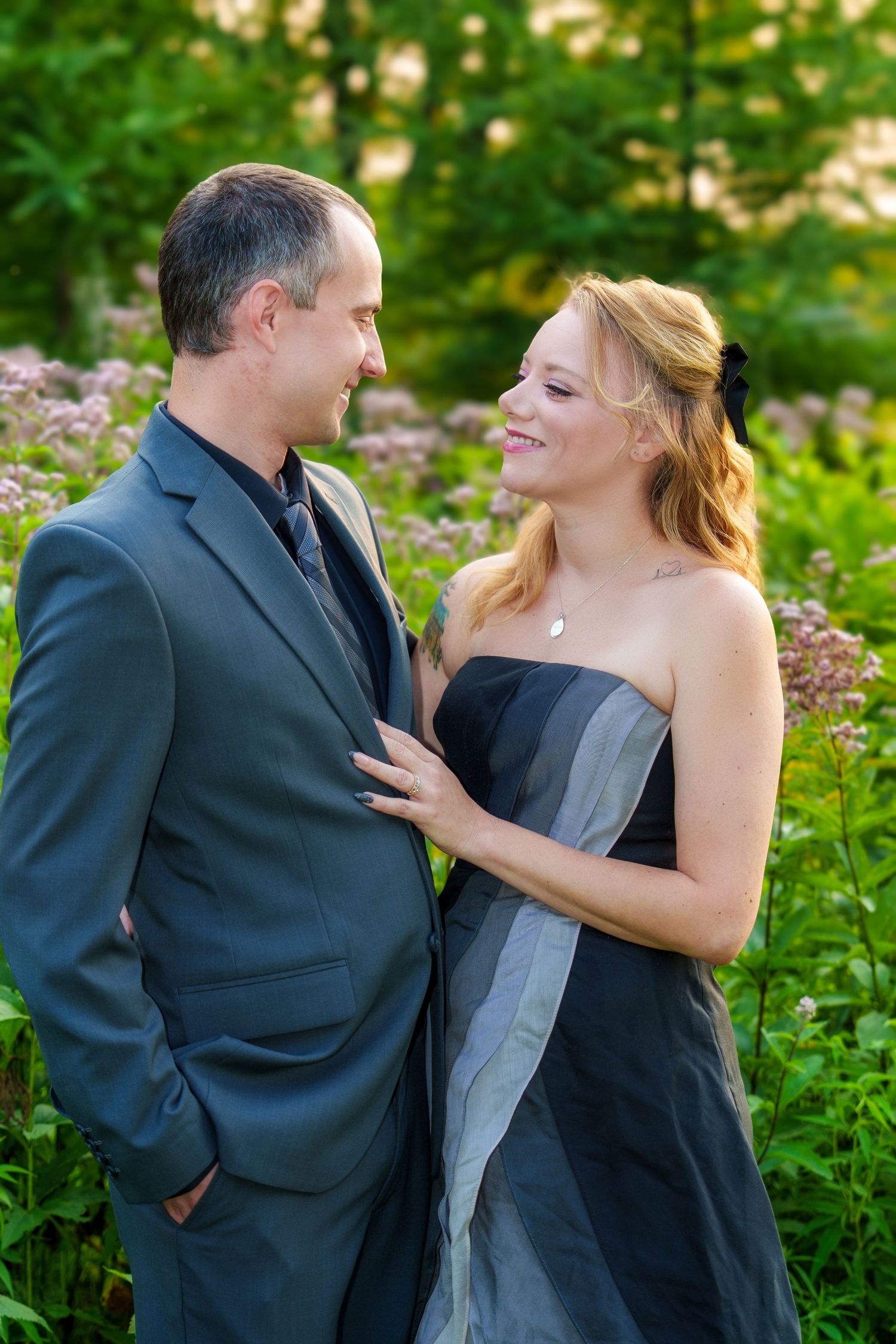 Couple in formal attire smiling at each other outdoors, woman in black strapless dress, man in gray suit, lush green background.
