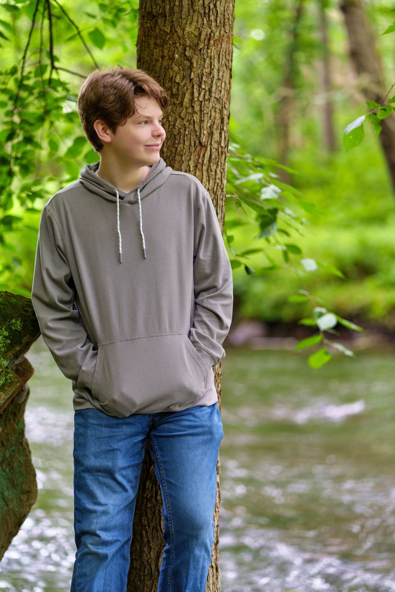 Teen boy in gray hoodie and jeans leaning against a tree in a lush green forest near a stream.