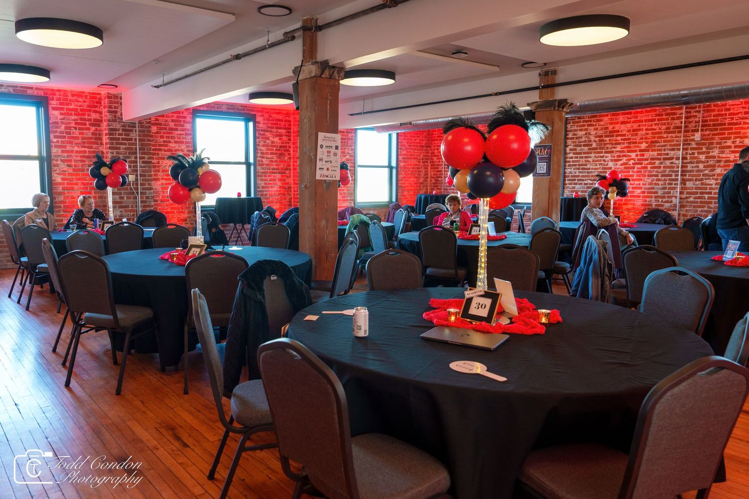 Elegant event venue with round tables, teal linens, black chairs, red and black balloon centerpieces, and exposed brick walls.
