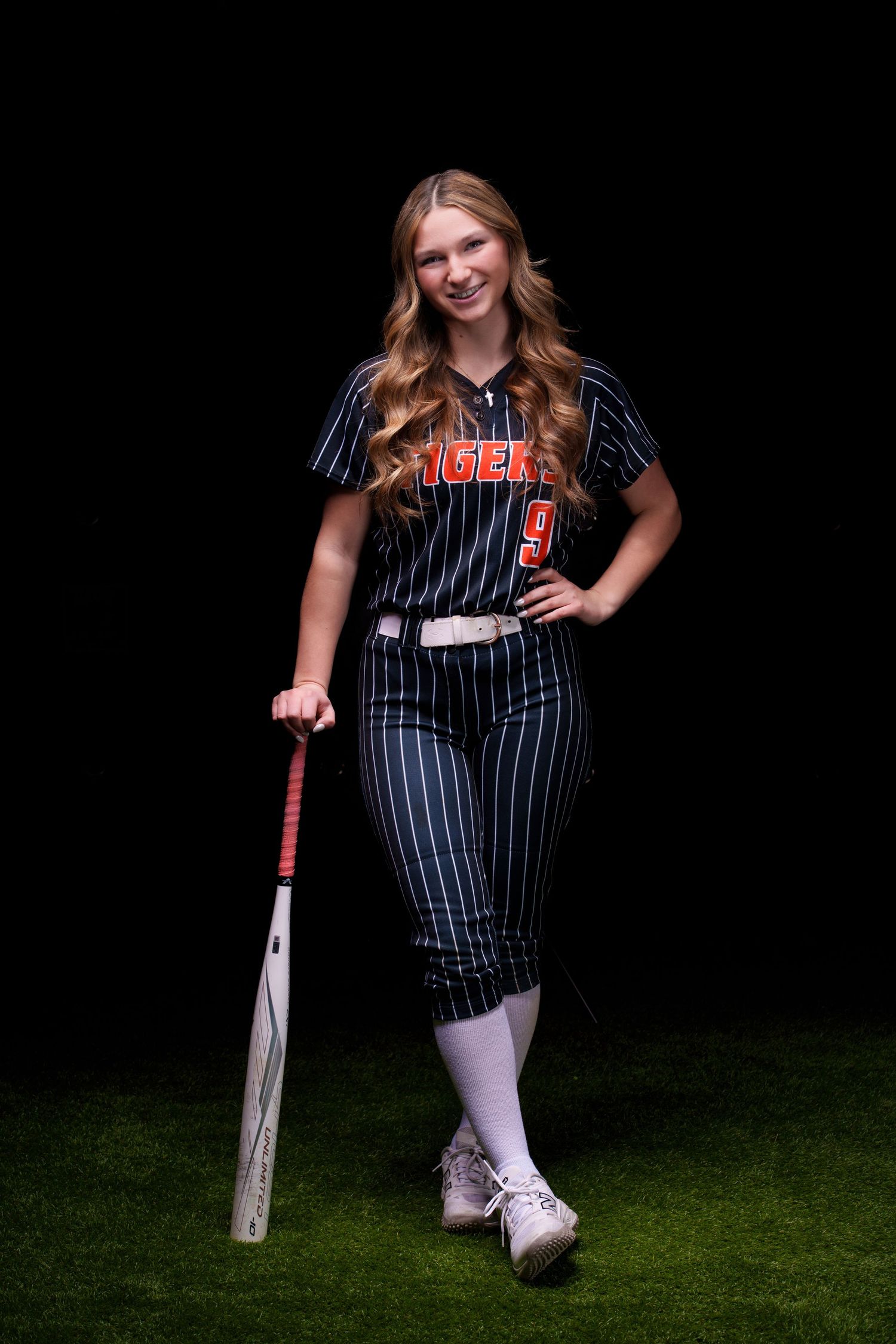 Young female softball player in Tigers uniform holding a bat, posing on grass against a dark background.