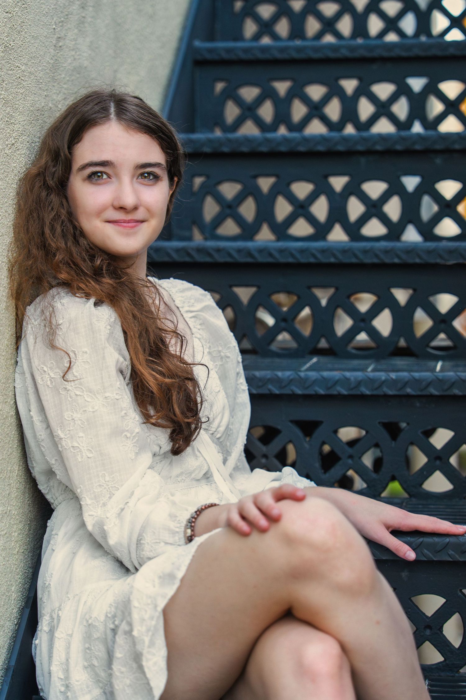 Young woman in white lace dress sitting on ornate blue metal stairs outdoors.