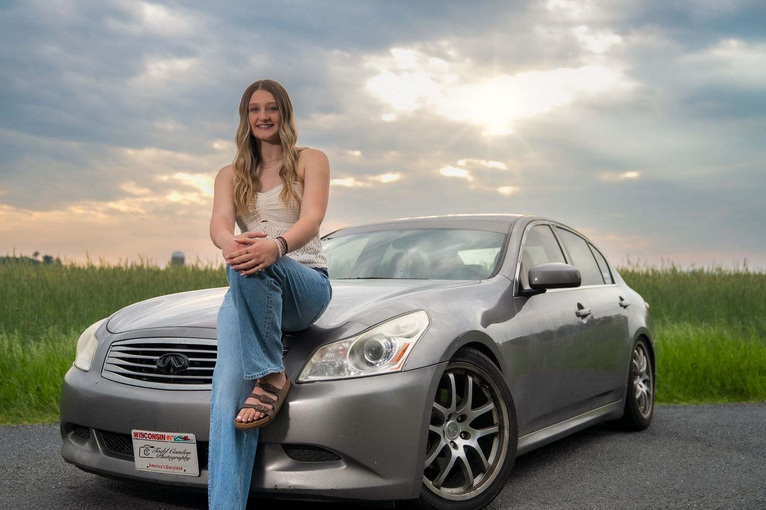 Silver luxury sedan car photographed in a rural setting with dramatic cloudy sky at sunset.