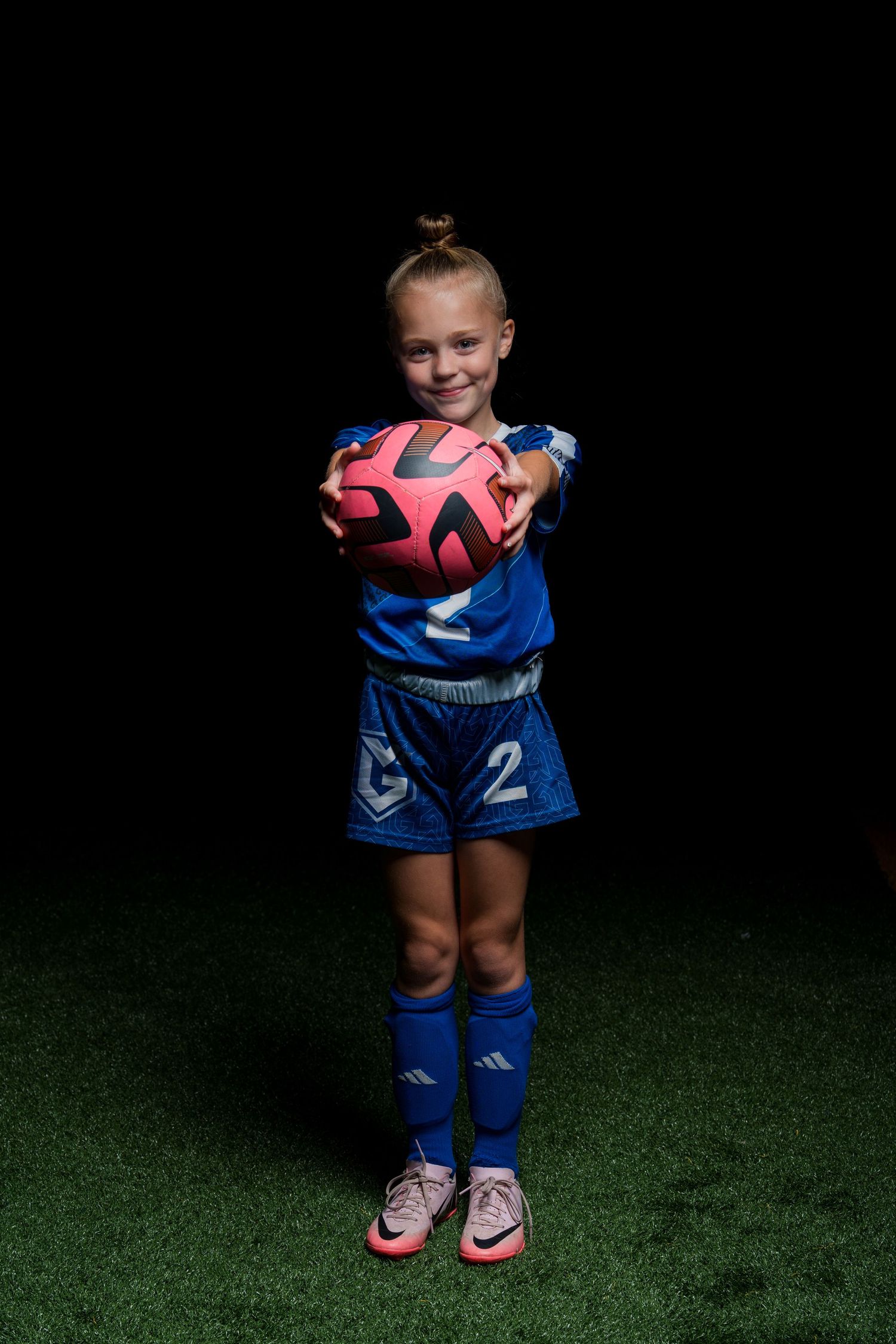 Young soccer player in blue uniform holds pink soccer ball against dark background.