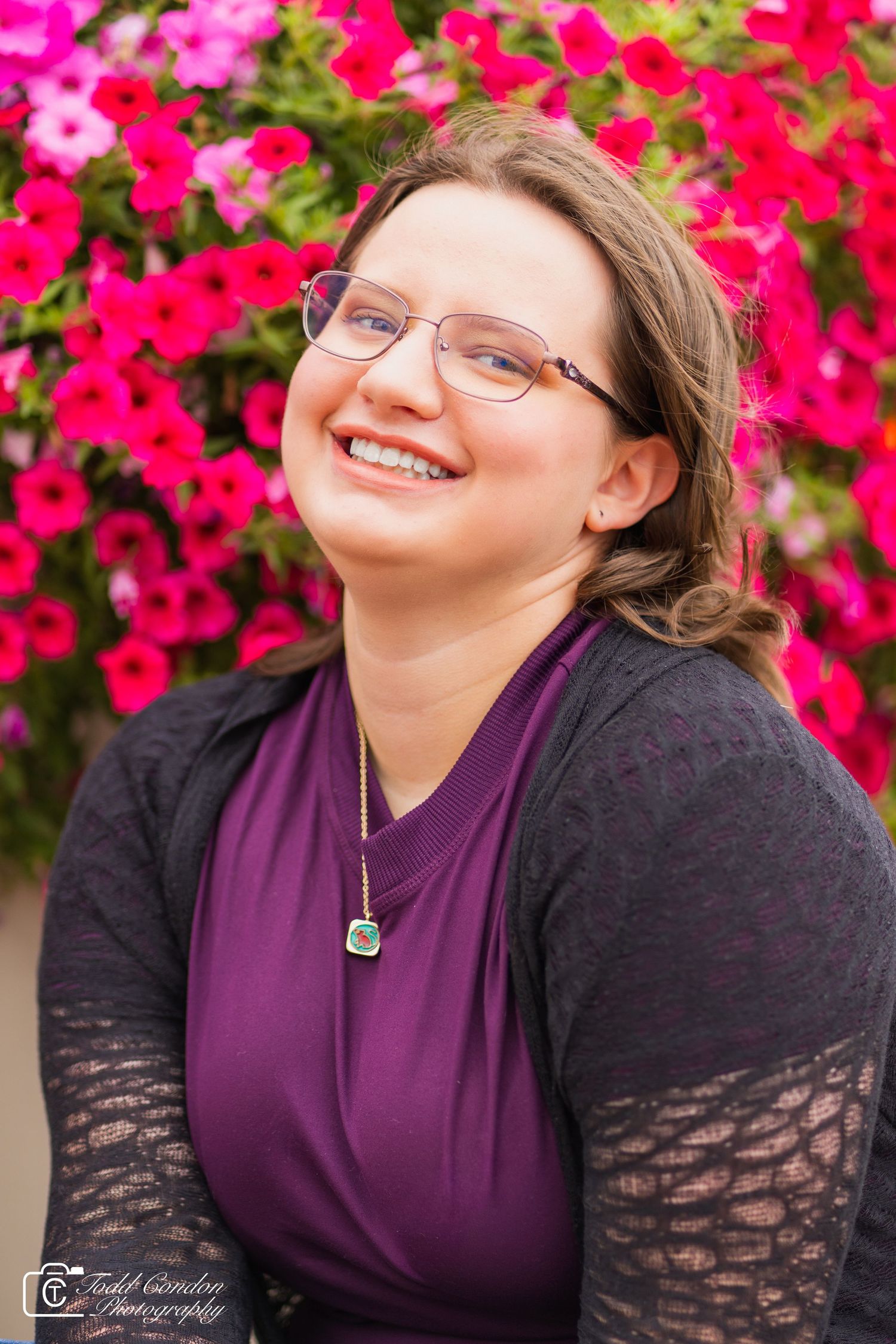A smiling figure in glasses and purple shirt poses against vibrant pink flowers in an outdoor portrait.