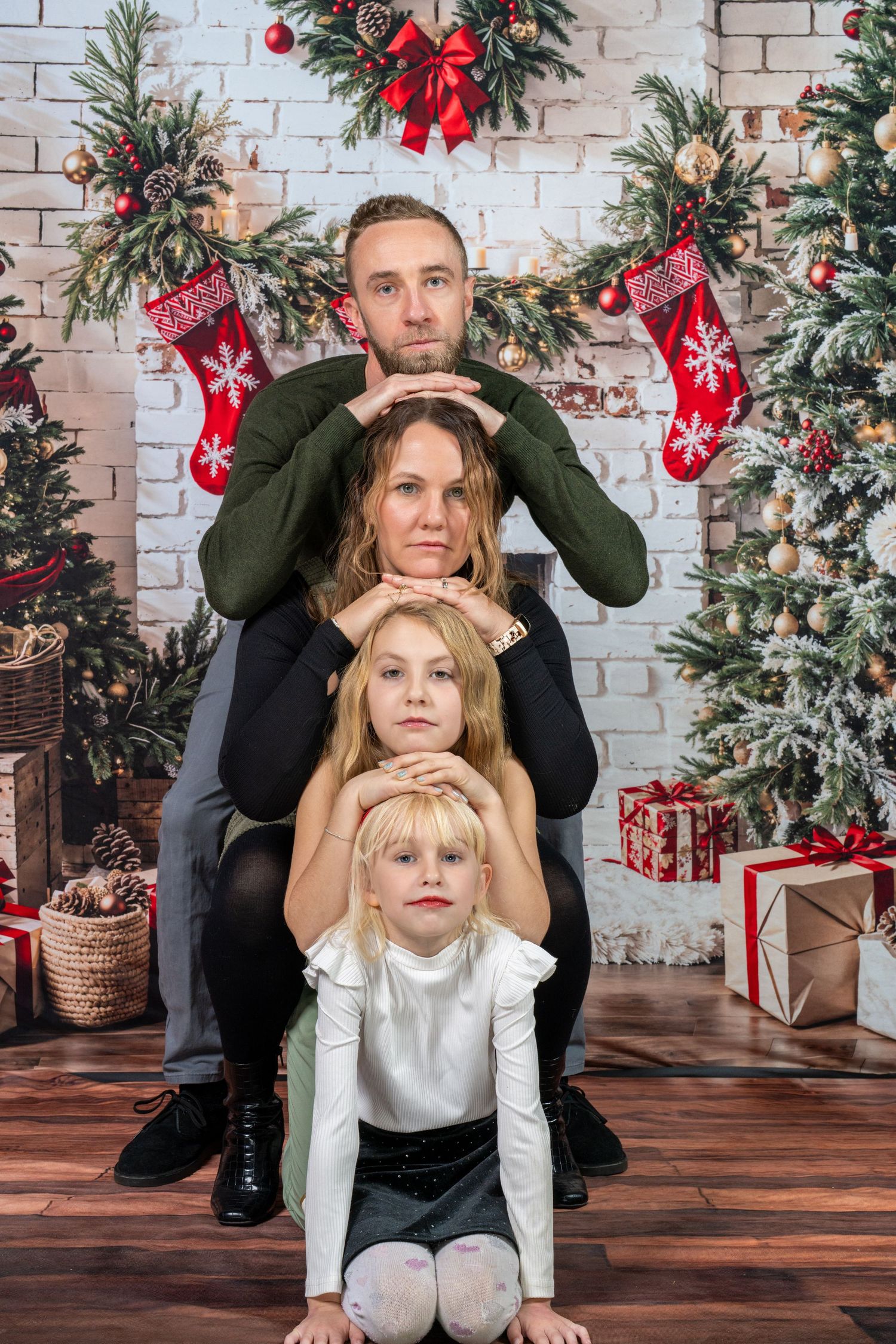 Family poses playfully in front of Christmas decorations with white brick wall and evergreen garland backdrop.