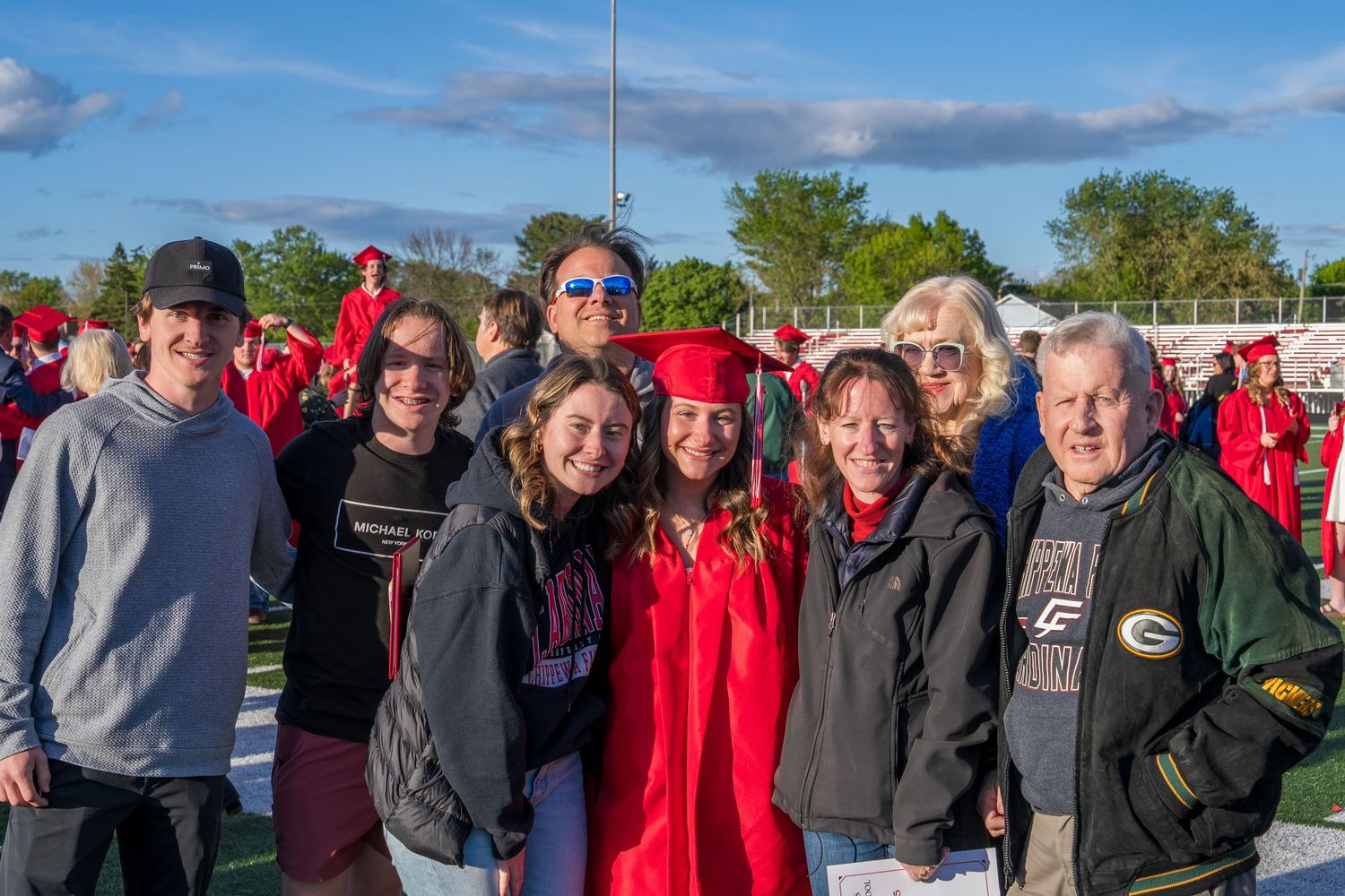 Group of family and friends posing with a high school graduate in red cap and gown at outdoor ceremony.