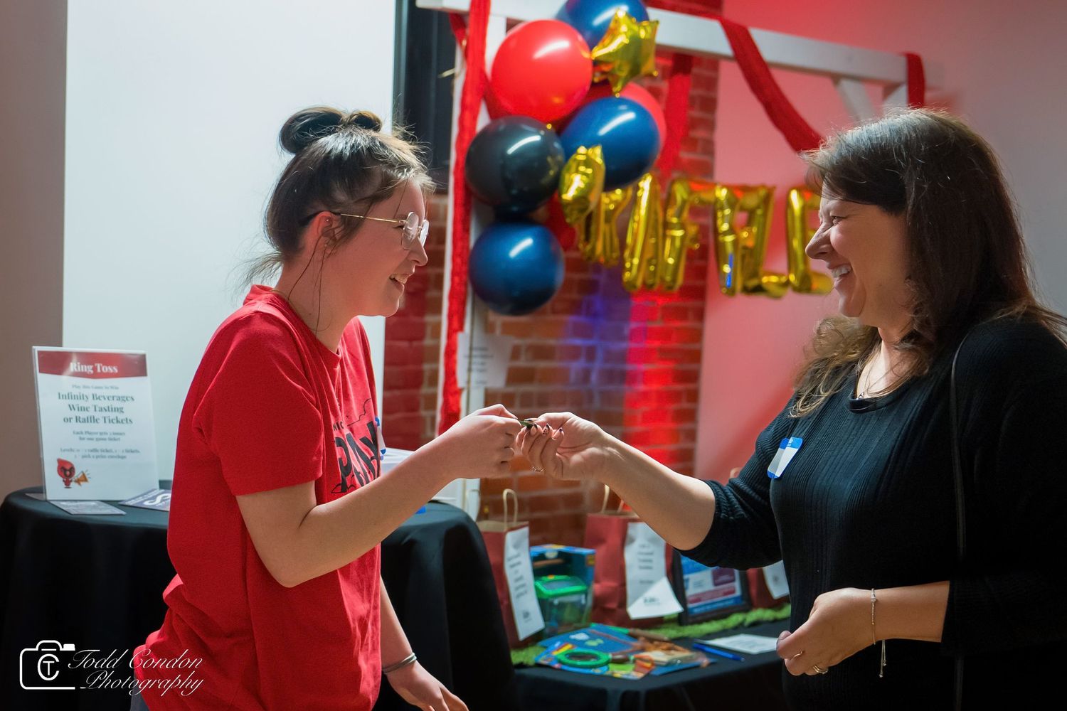 Two women exchanging items at a festive event with colorful balloons and decorations in the background.