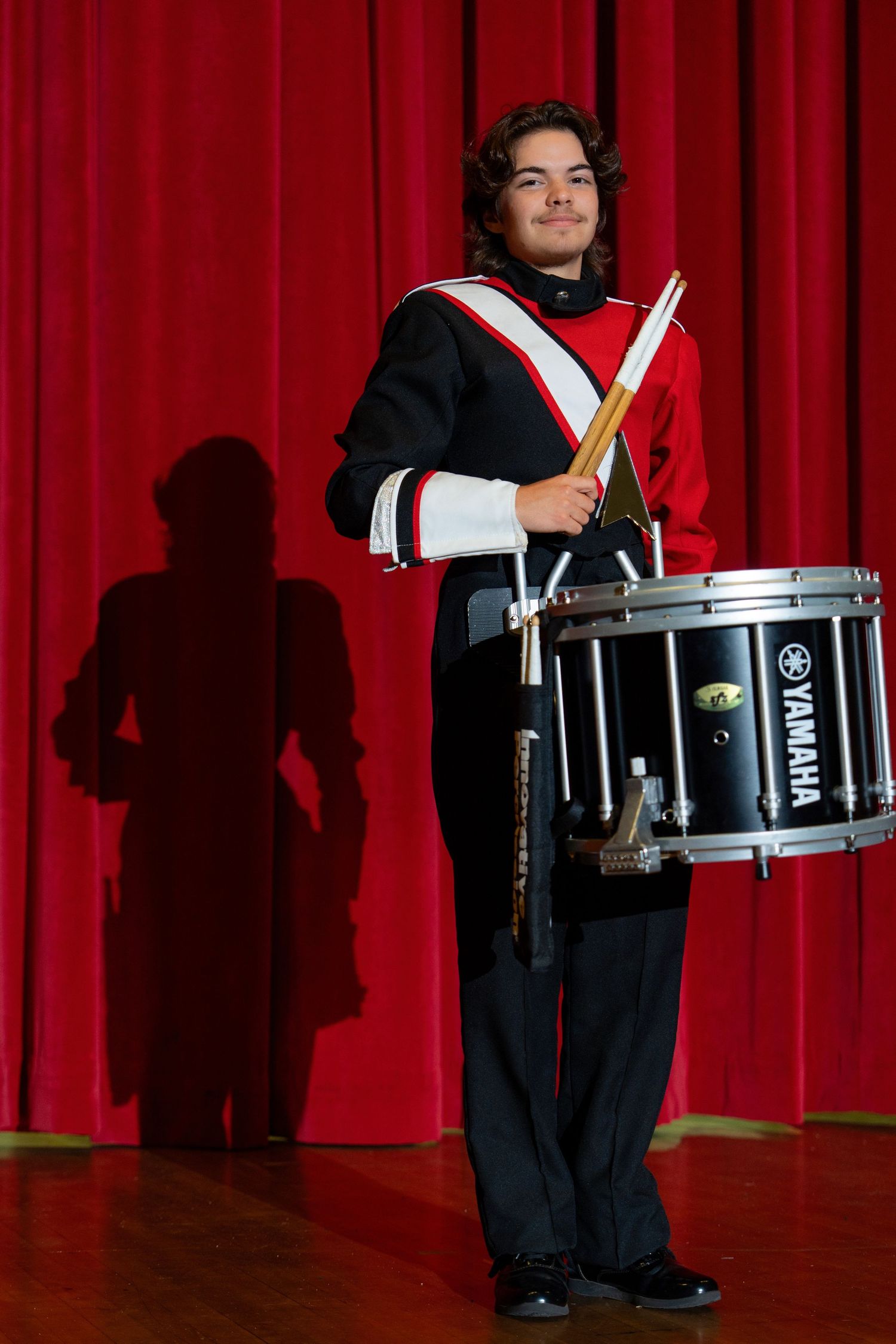 Drummer in marching band uniform stands with drumsticks and Yamaha snare drum against red curtain backdrop.