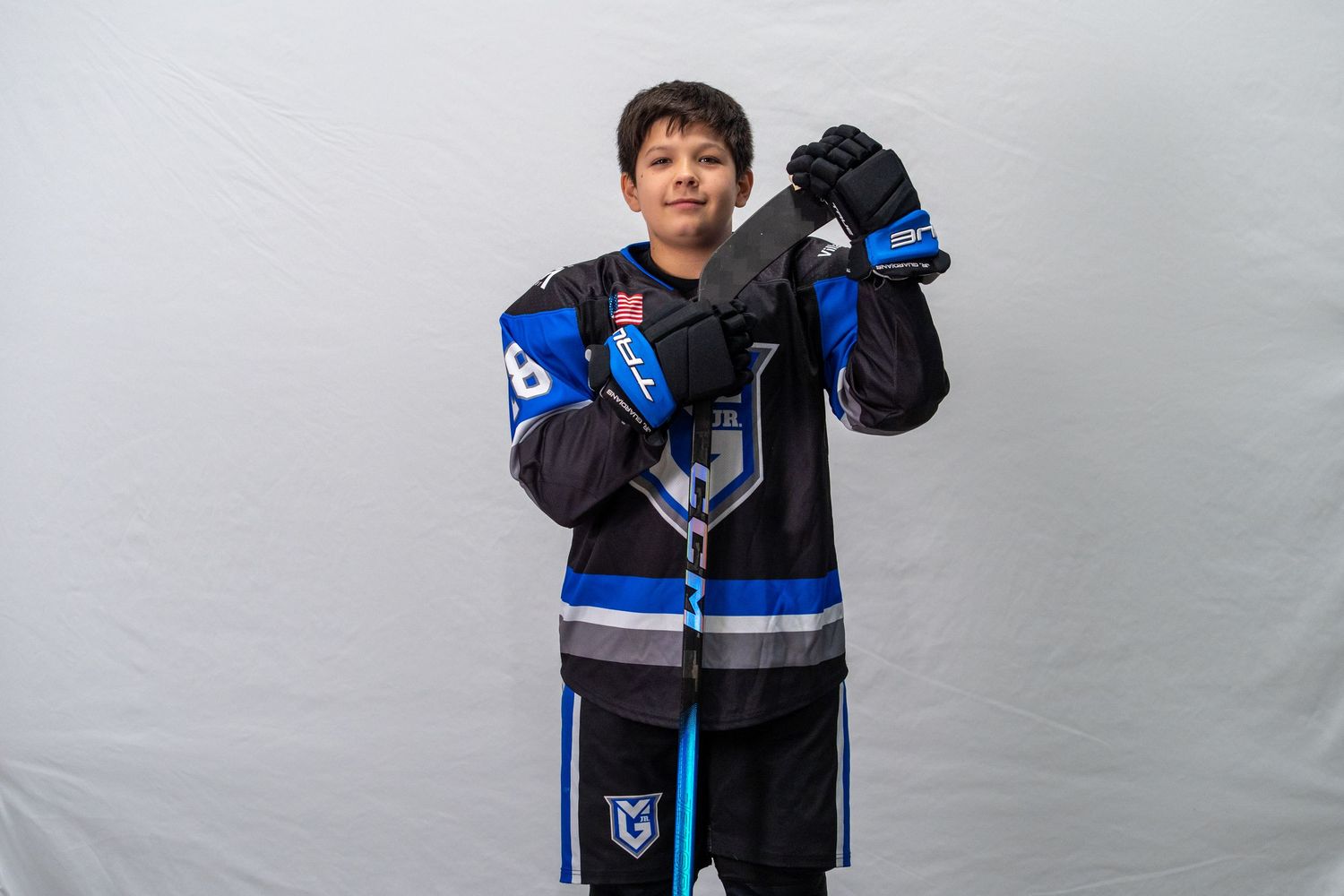 Young hockey player in blue and black jersey poses with stick and gloves against white background.
