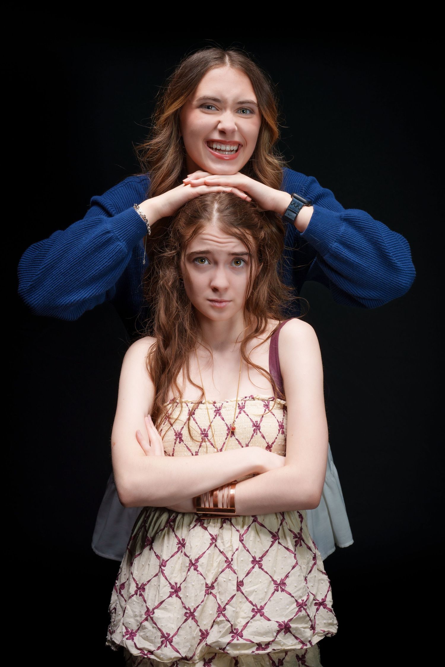 Two young women posing on black background, one smiling resting hands on other's head who stands arms crossed.