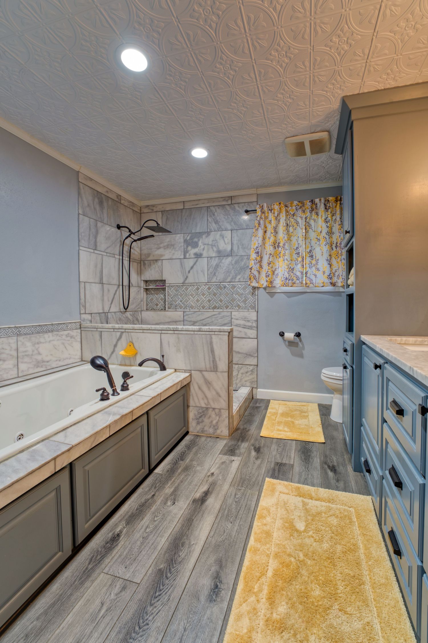 Spacious bathroom with gray cabinets, soaking tub, tiled walk-in shower, hardwood floors, and yellow accent rugs.