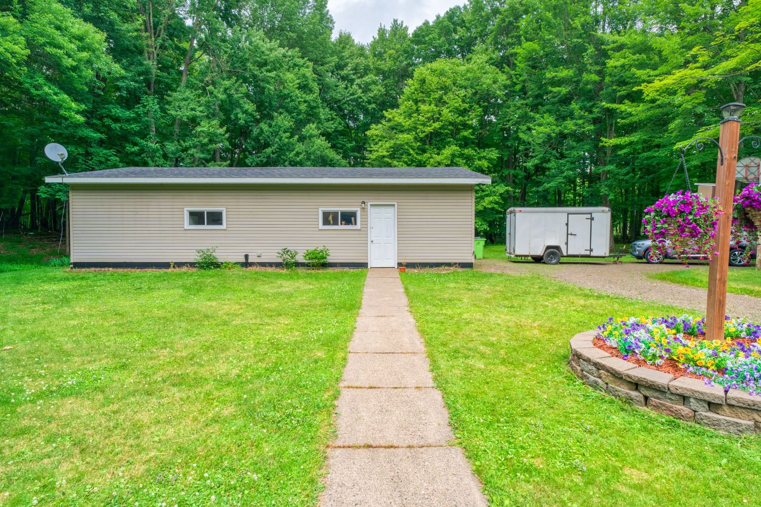 Ranch-style home with tan siding, white door, concrete path, manicured lawn, flower garden, and lush green trees in background.