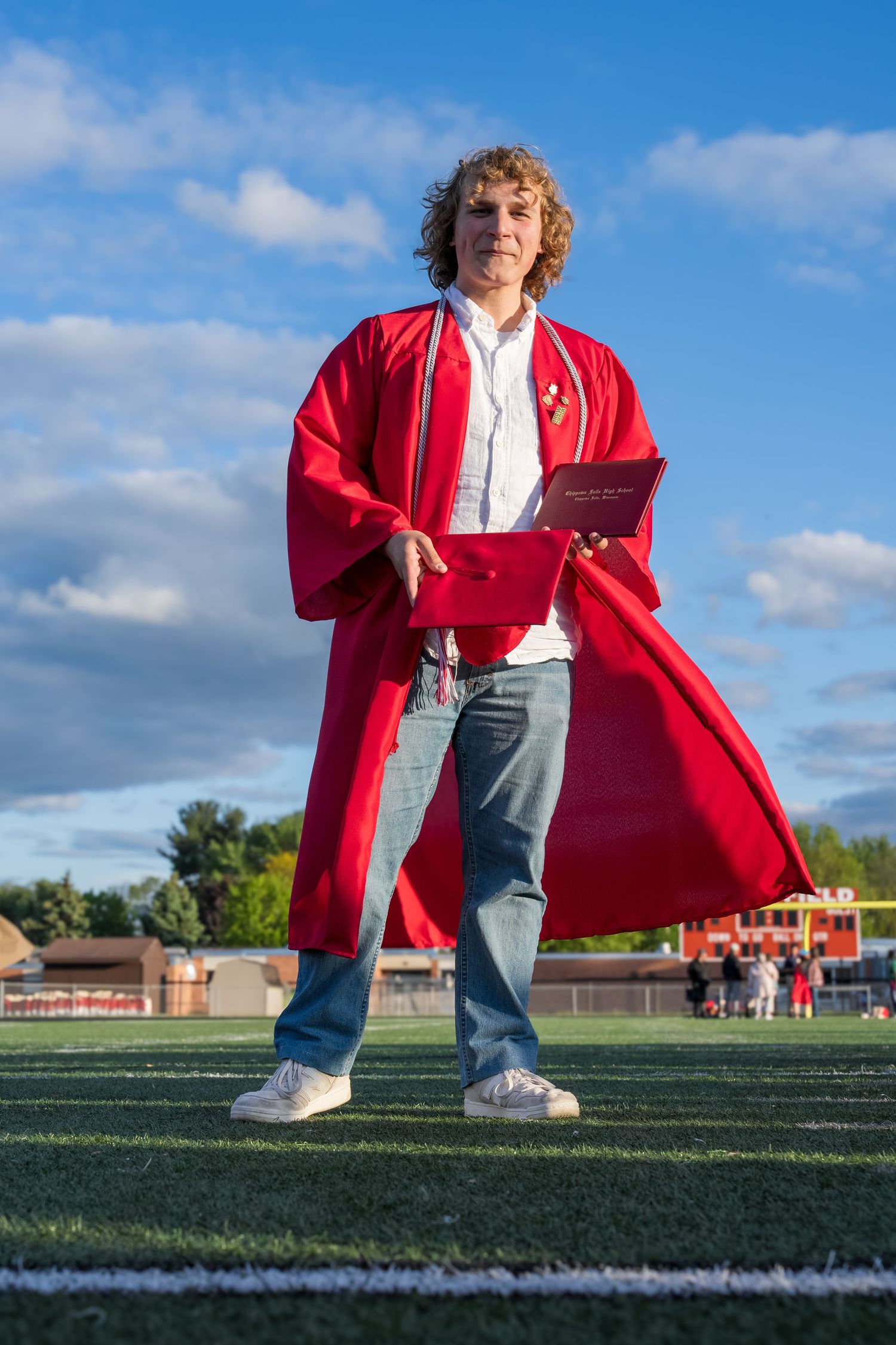 Young man in red graduation gown holding diploma on football field under blue sky.