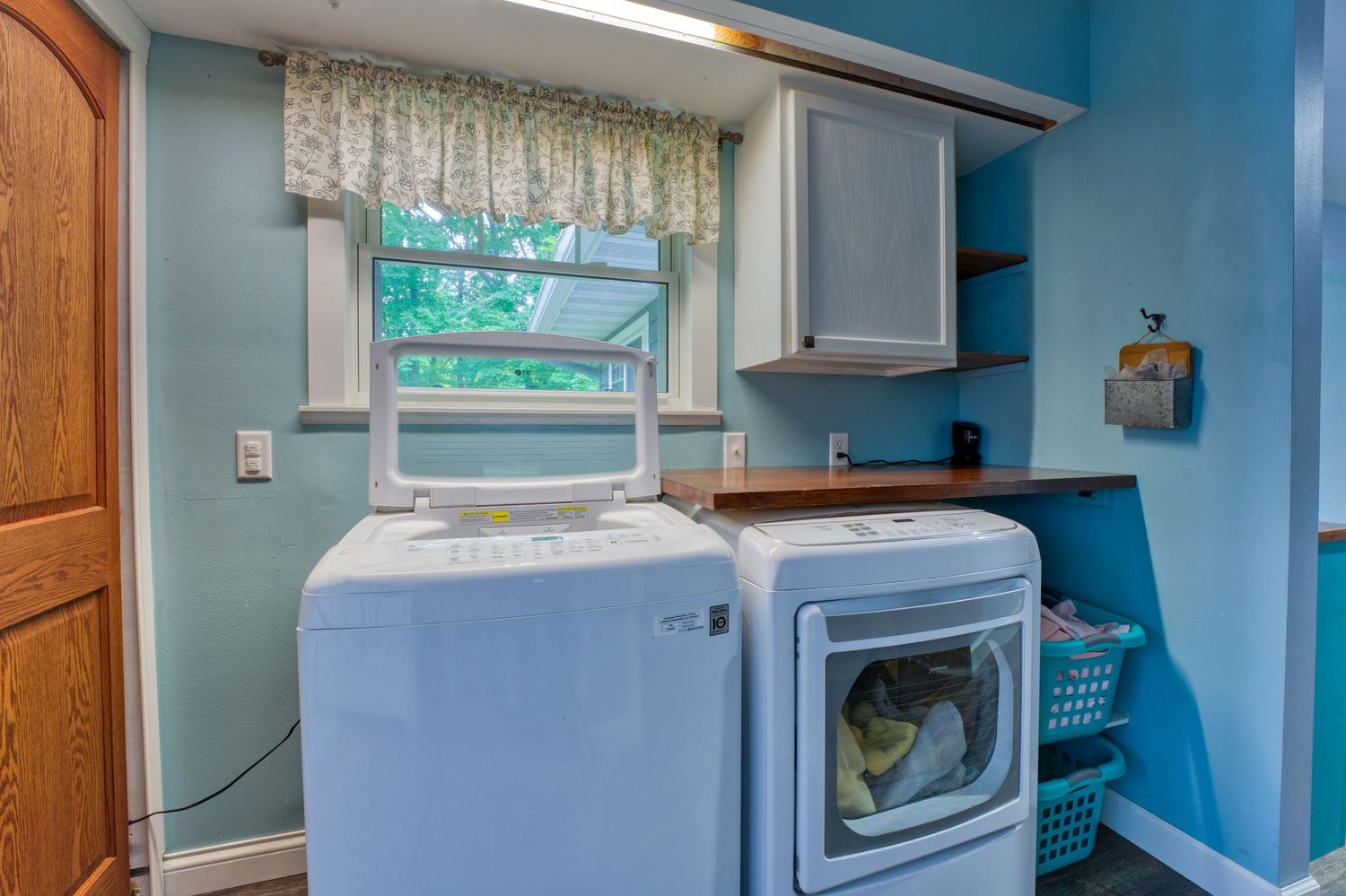 Laundry room with white washer and dryer, blue walls, wood countertop, cabinet storage, and a window with floral valance.