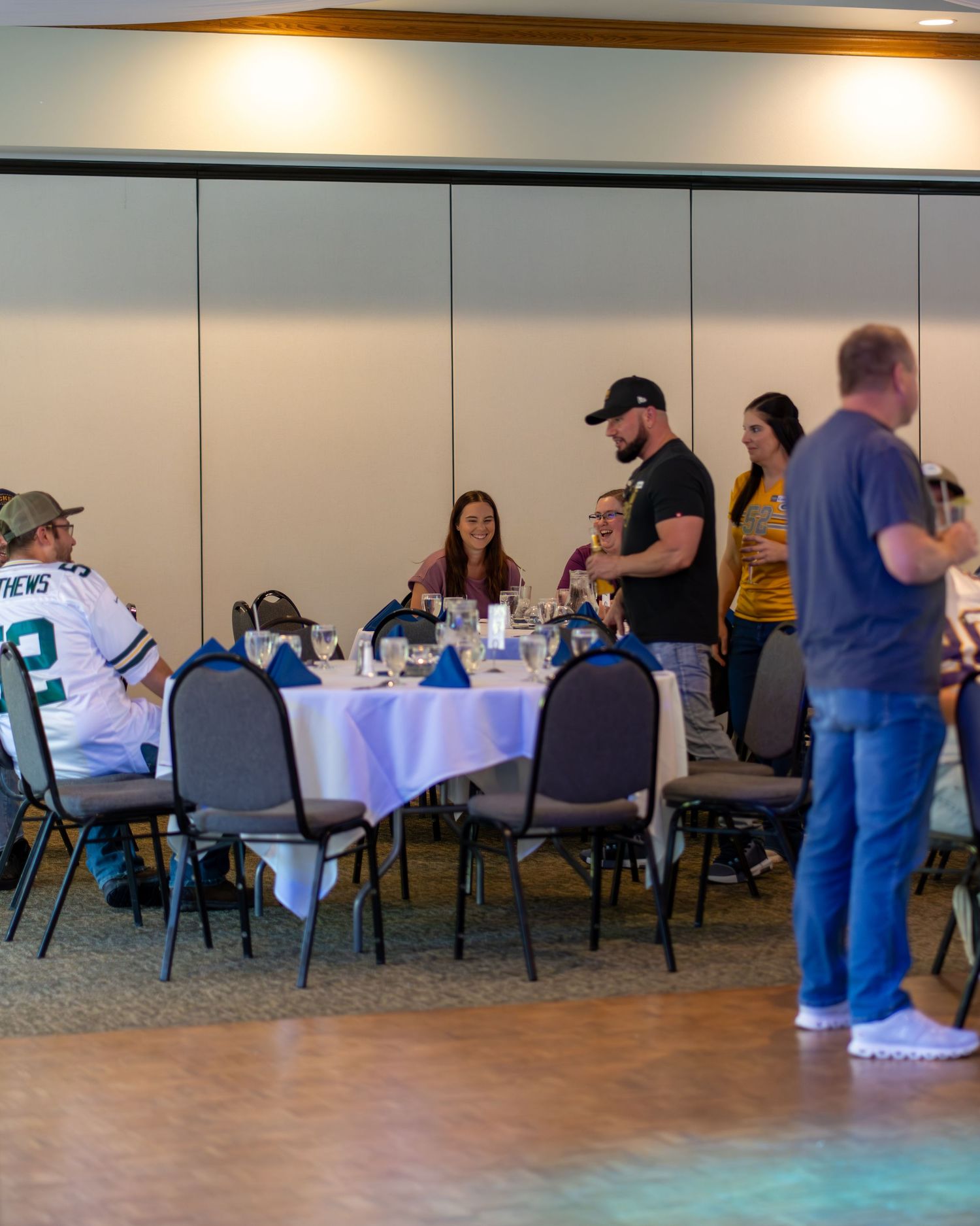 People gather at round tables with white tablecloths in a banquet hall setting during a social event.
