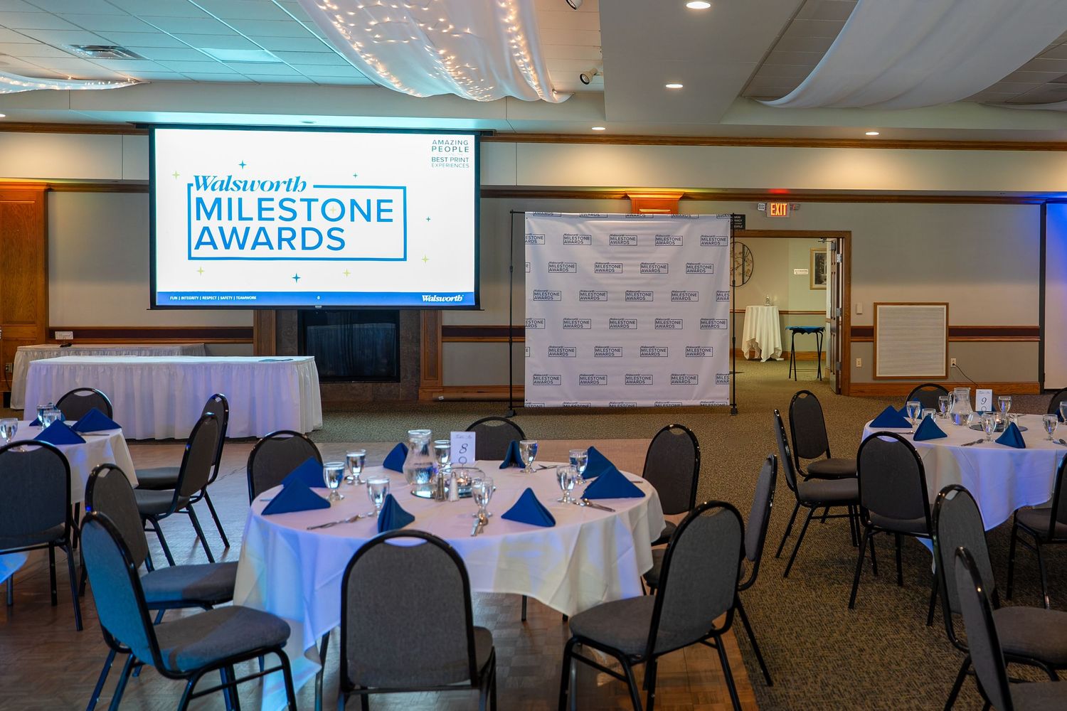 A banquet room setup with round tables, blue tablecloths and a projection screen displaying Milestone Awards presentation.