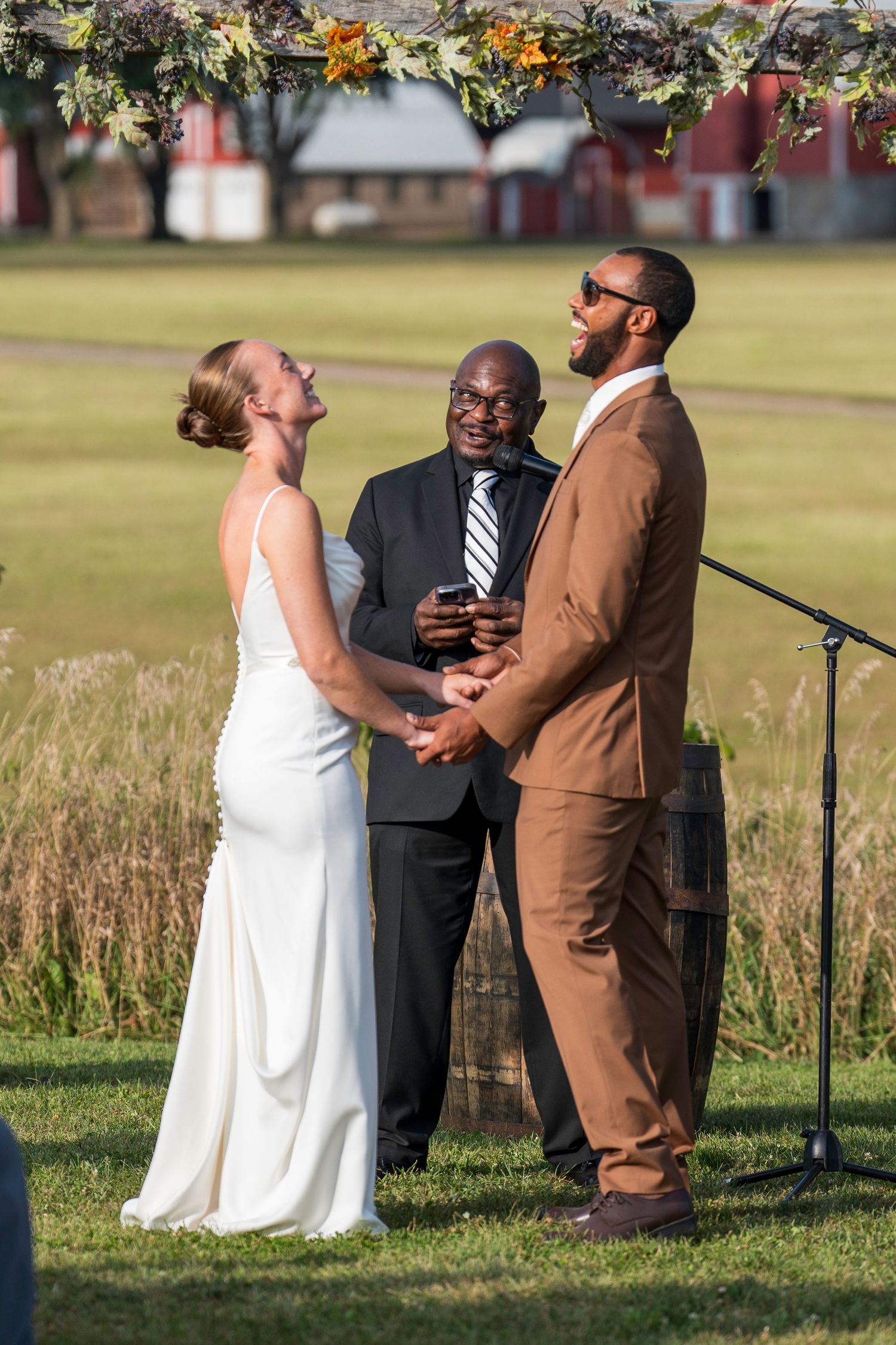 A wedding ceremony takes place outdoors on a sunny day with a couple and officiant standing in tall grass.