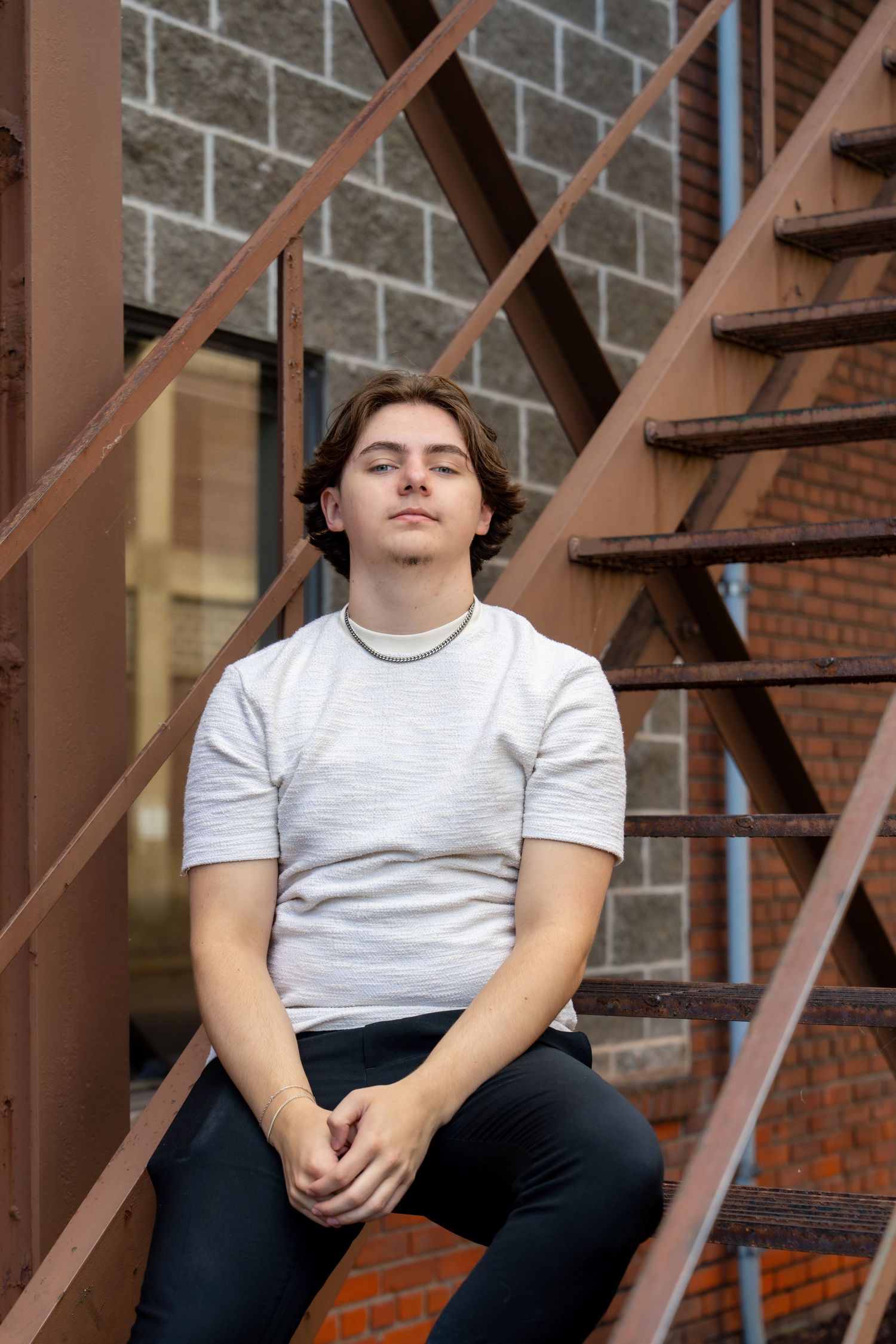A person in a white t-shirt sits casually on metal stairs against a brick wall backdrop.