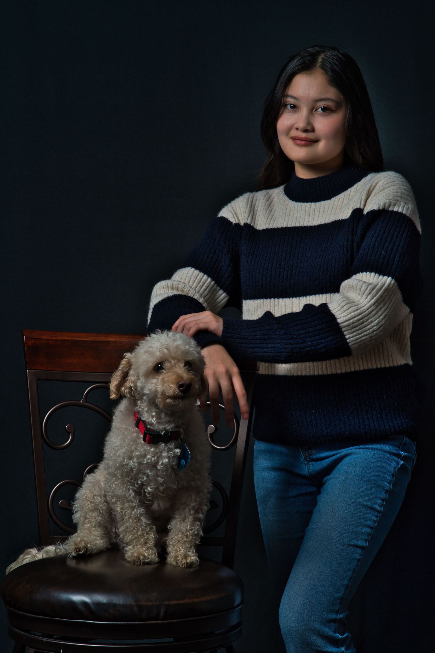 A person in a striped sweater sits with their small gray poodle mix dog on a dark leather chair against a black background.