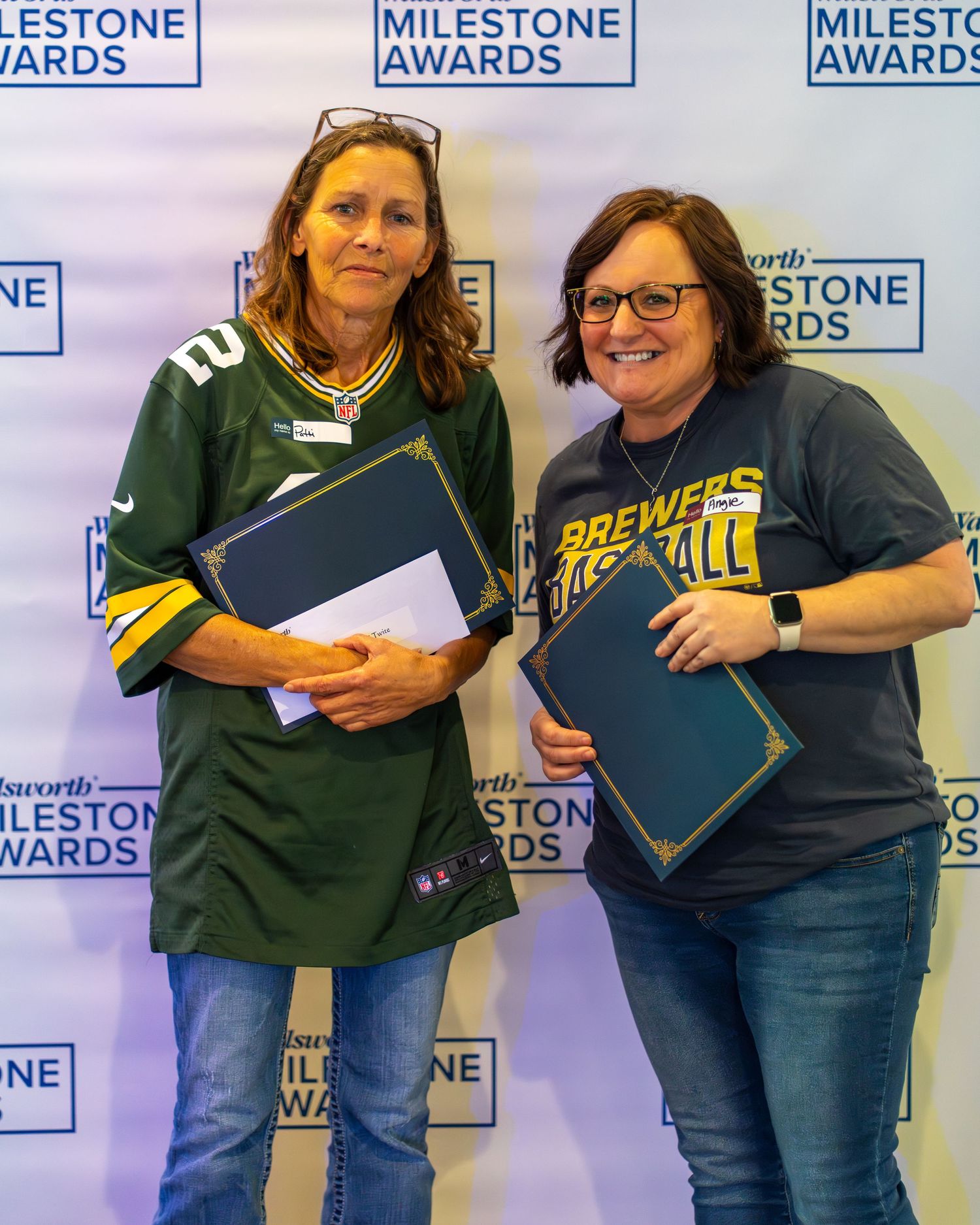 Two people in casual attire pose together holding award certificates against a branded backdrop with Milestone Awards logos.