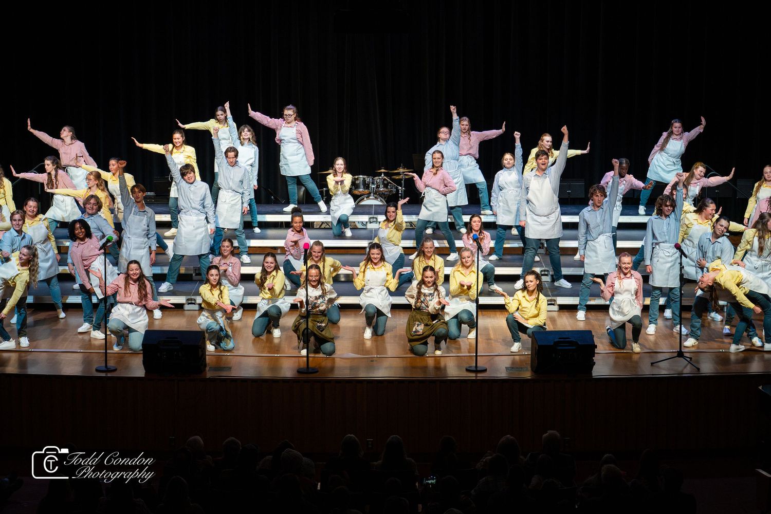 Performers in white outfits dance energetically across a dark stage during a theatrical production.