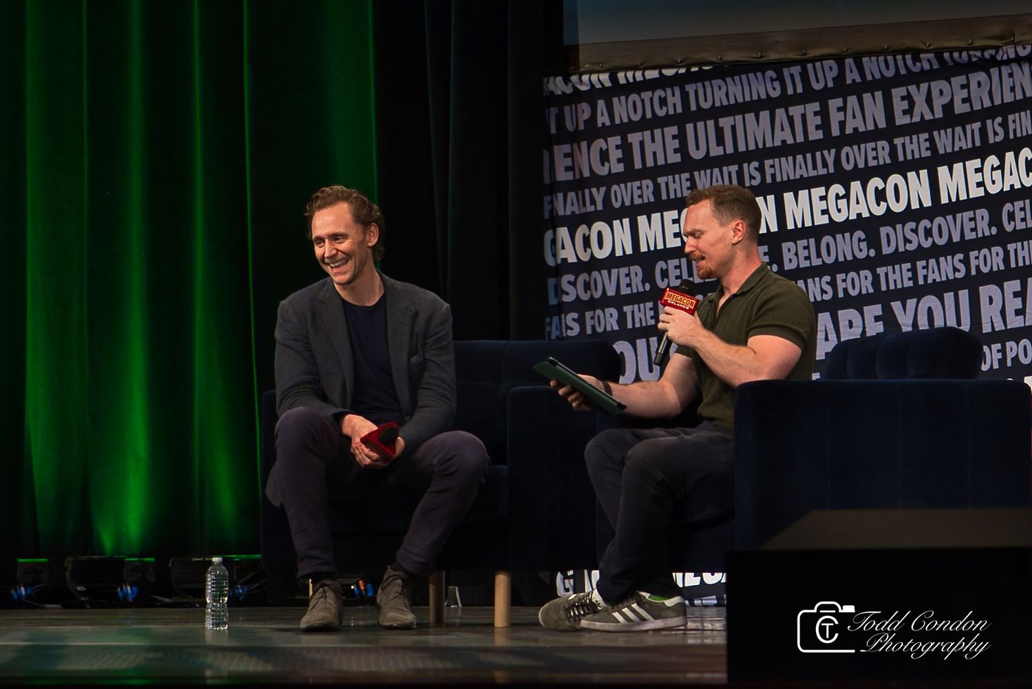 Two people sit on stage at a conference event with green lighting and text banners visible in the background.