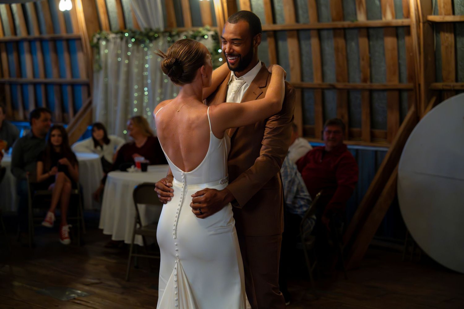 A couple shares their first dance at a rustic barn wedding reception with guests watching in the background.
