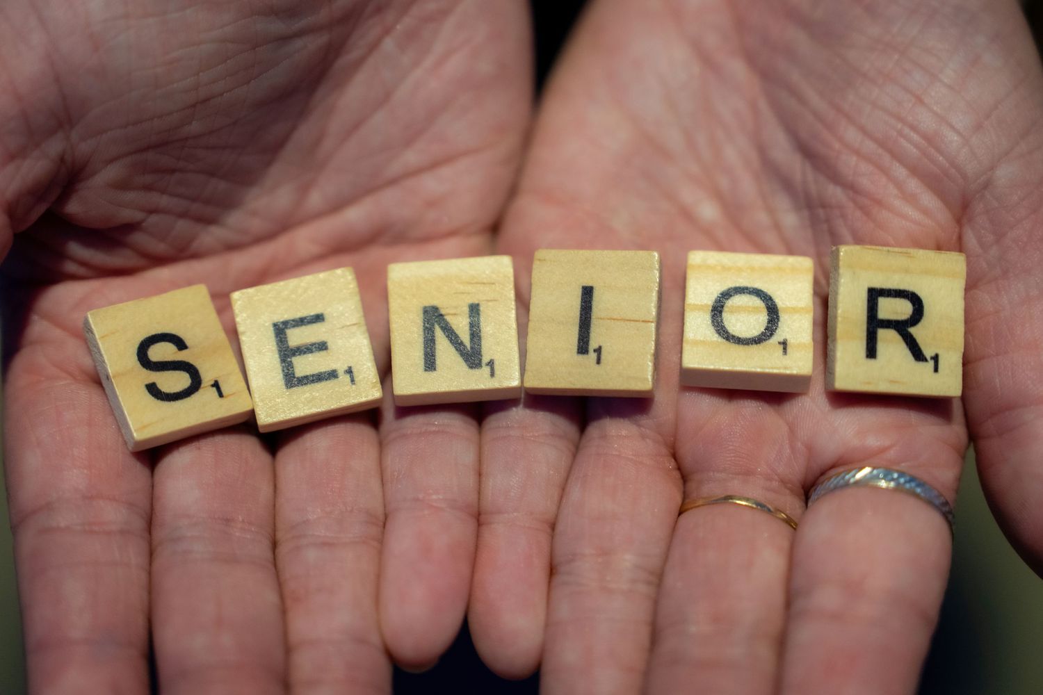 Scrabble letter tiles spell out the word 'SENIOR' held up by multiple hands against a blurred background.