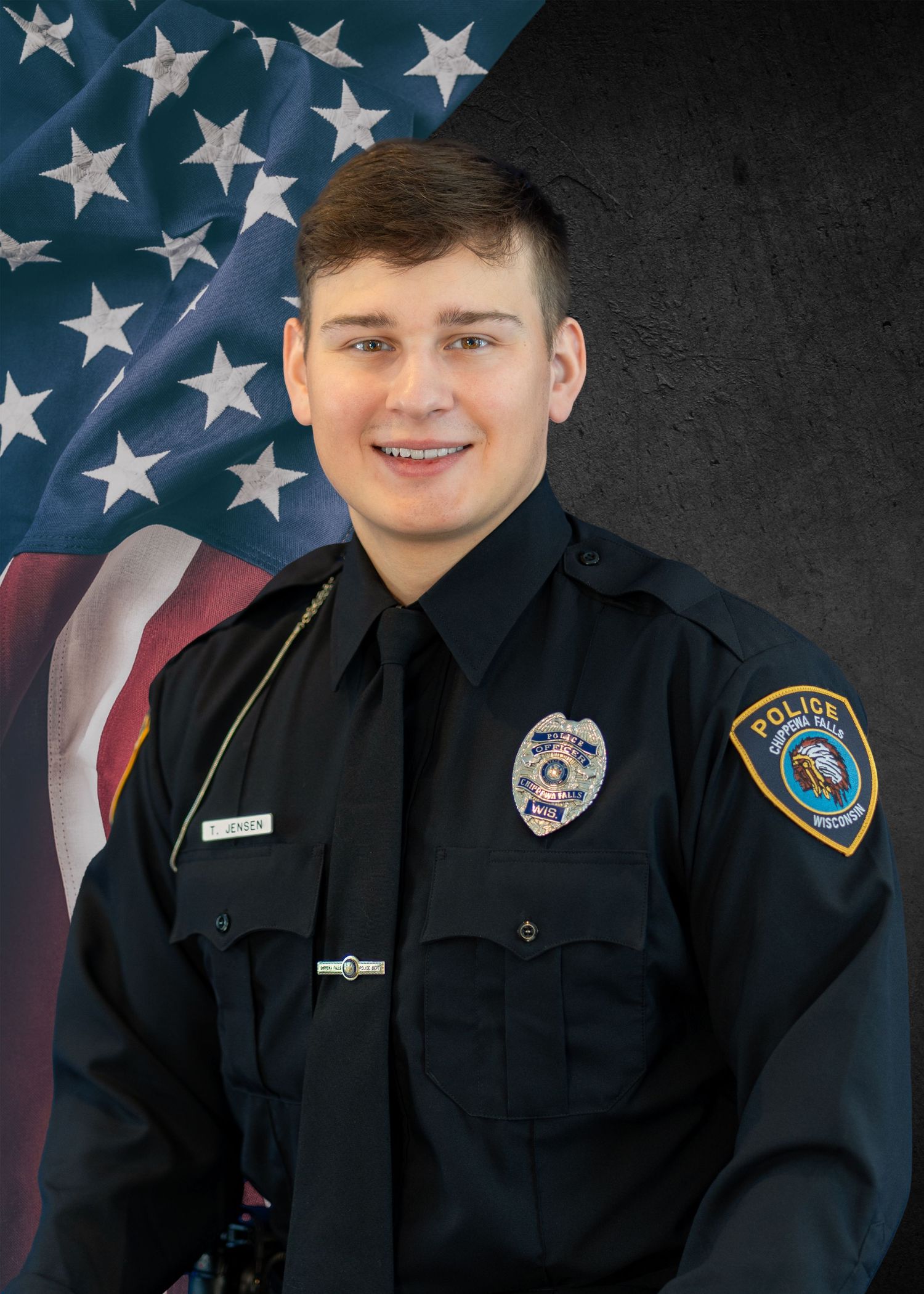 Young male police officer in black uniform smiling before American flag background in official portrait.