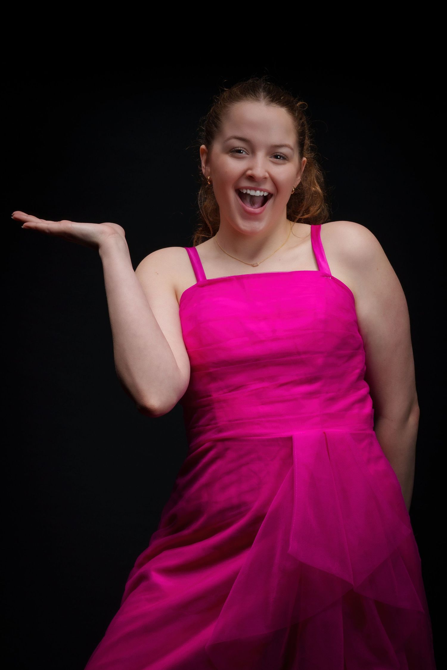Young woman in bright pink formal dress smiling with hand raised, posing against black background.