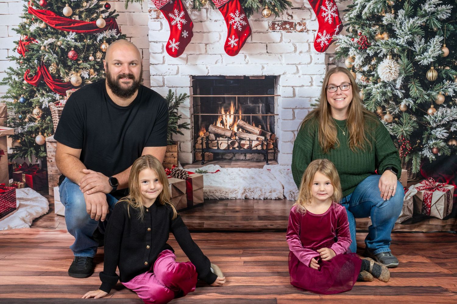 Family poses in front of white brick wall decorated with red stockings and holiday greenery for Christmas photos.